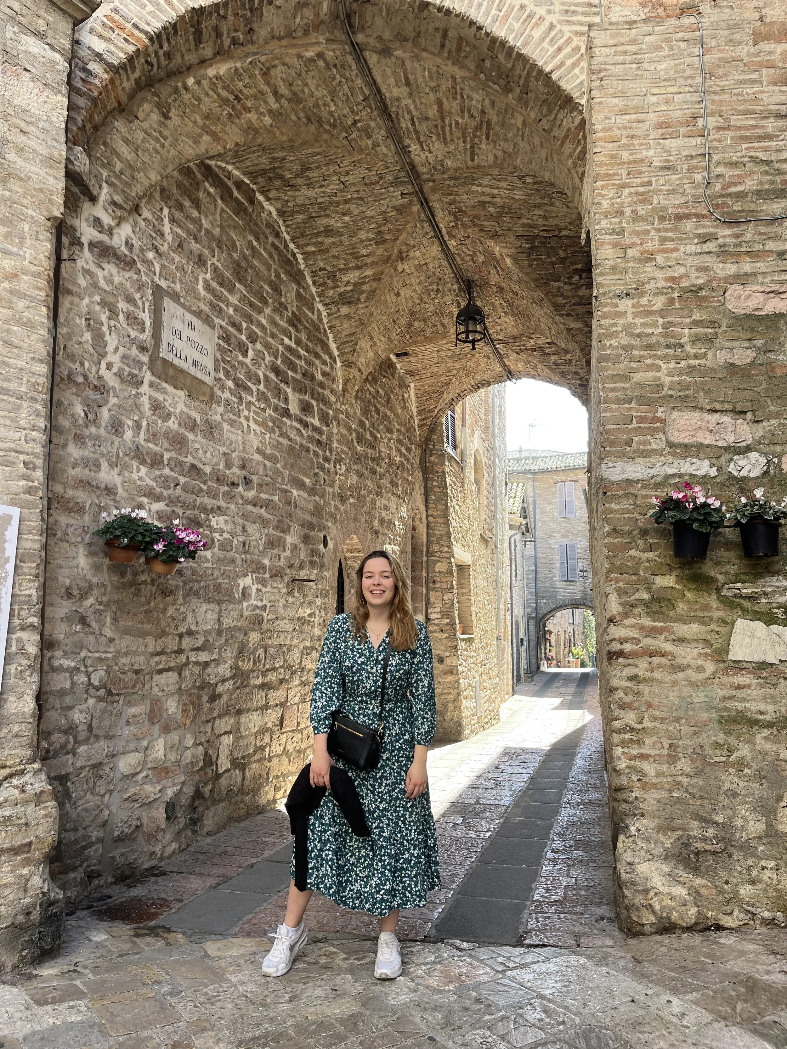 A woman standing in an old stone alleyway with an arch overhead, smiling at the camera, wearing a patterned dress, white sneakers, and carrying a black purse.