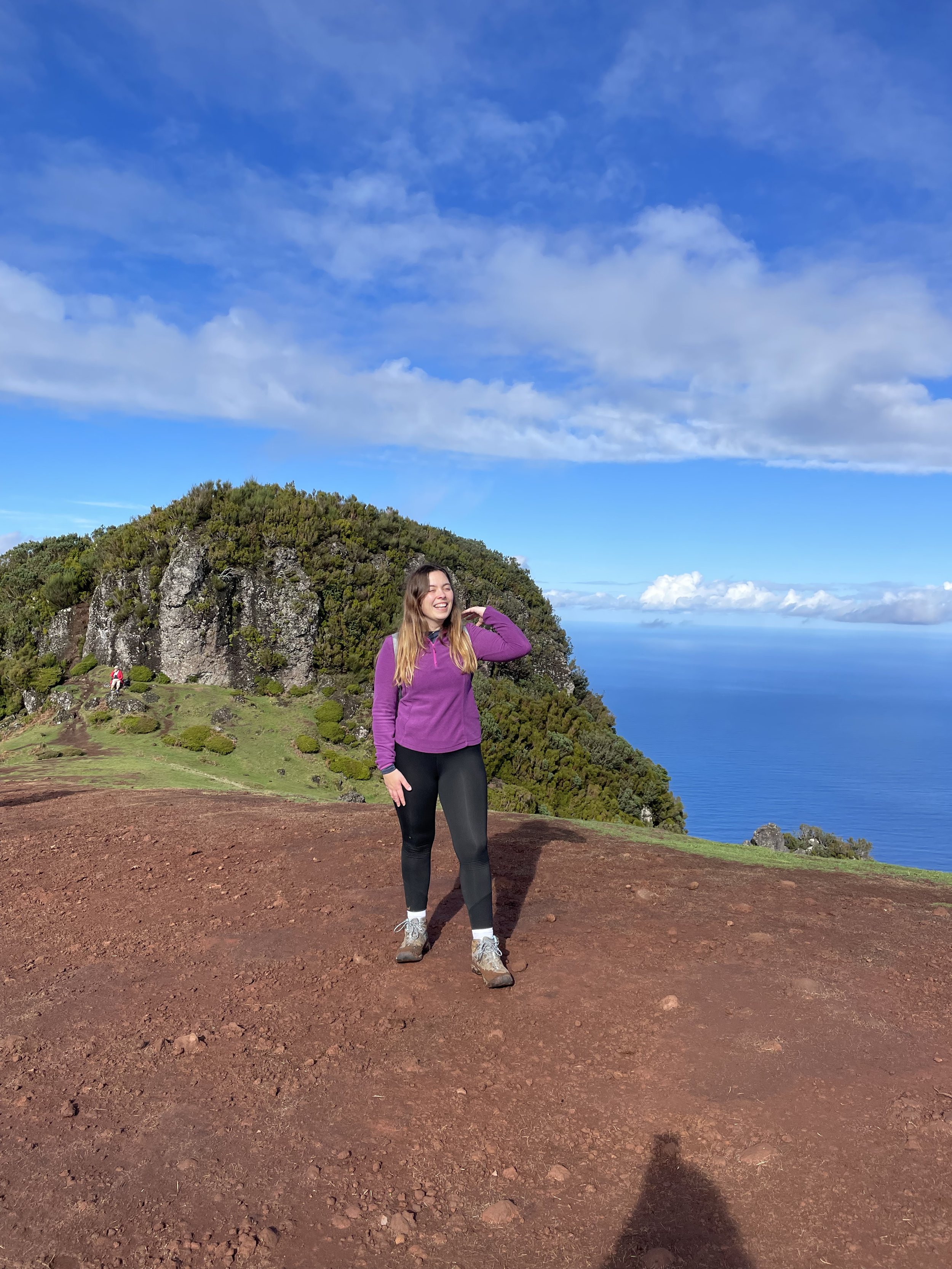 A woman in purple jacket and black pants standing on reddish dirt ground with a green hill and cliffs in the background under a partly cloudy sky near the ocean.