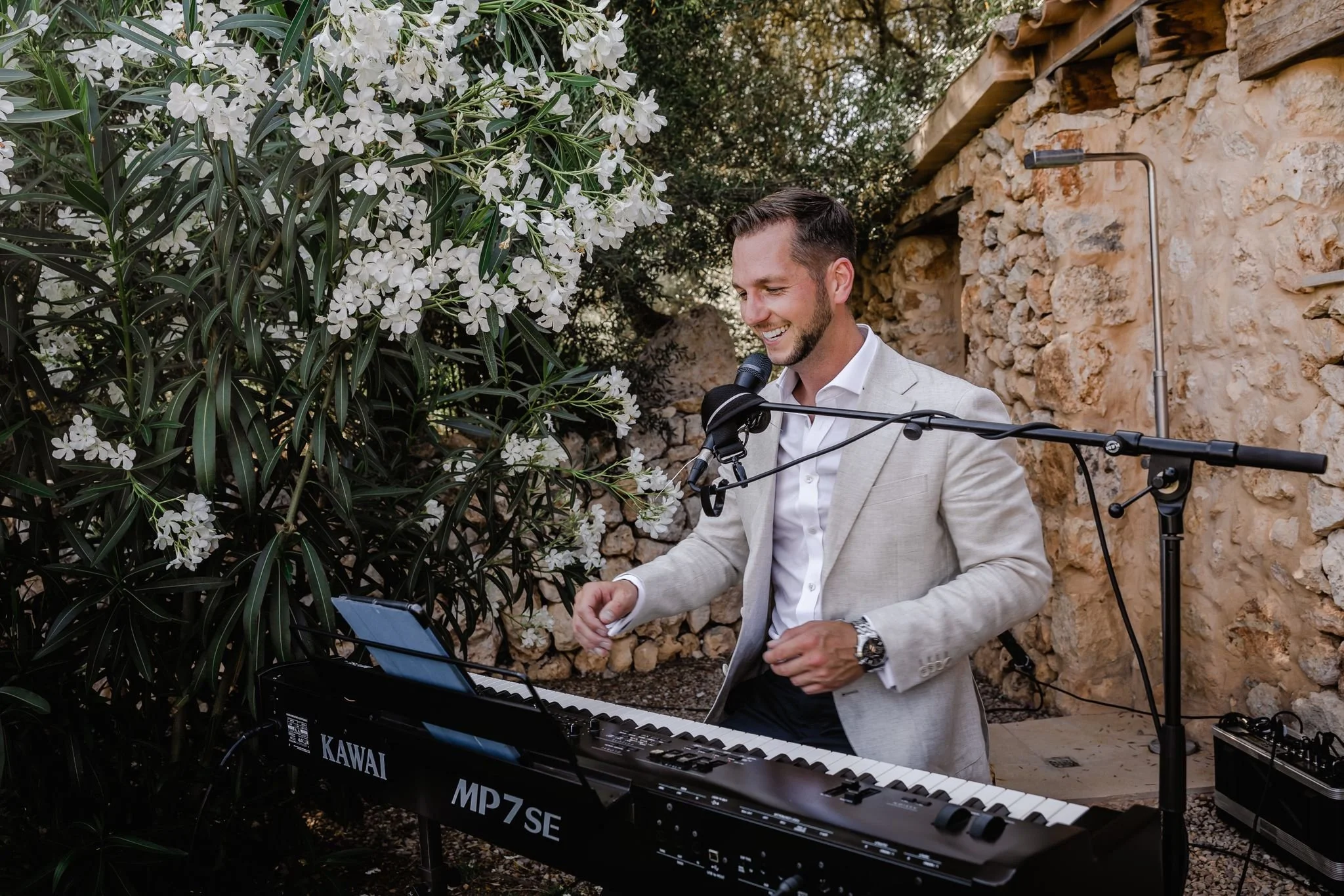 A man in a light-colored suit playing a keyboard and singing into a microphone outdoors next to flowering bushes and a stone wall.