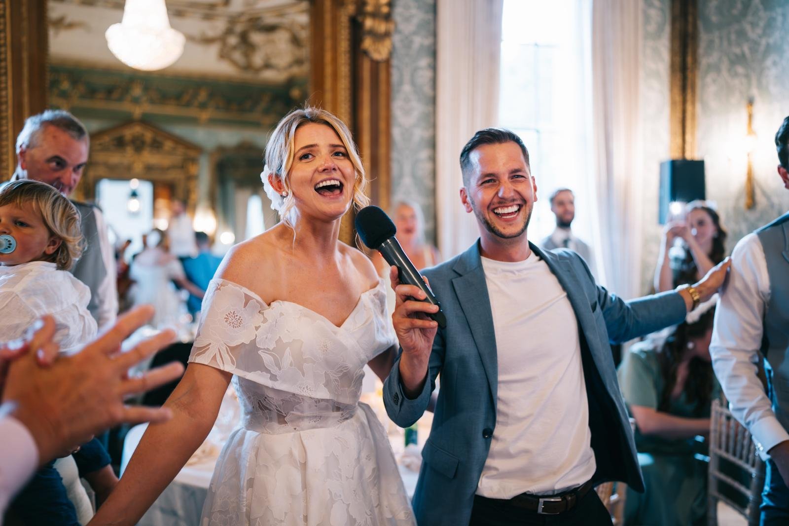 A happy woman in a white dress holding a microphone, standing next to a smiling man in a suit jacket, at a wedding reception with guests in the background.