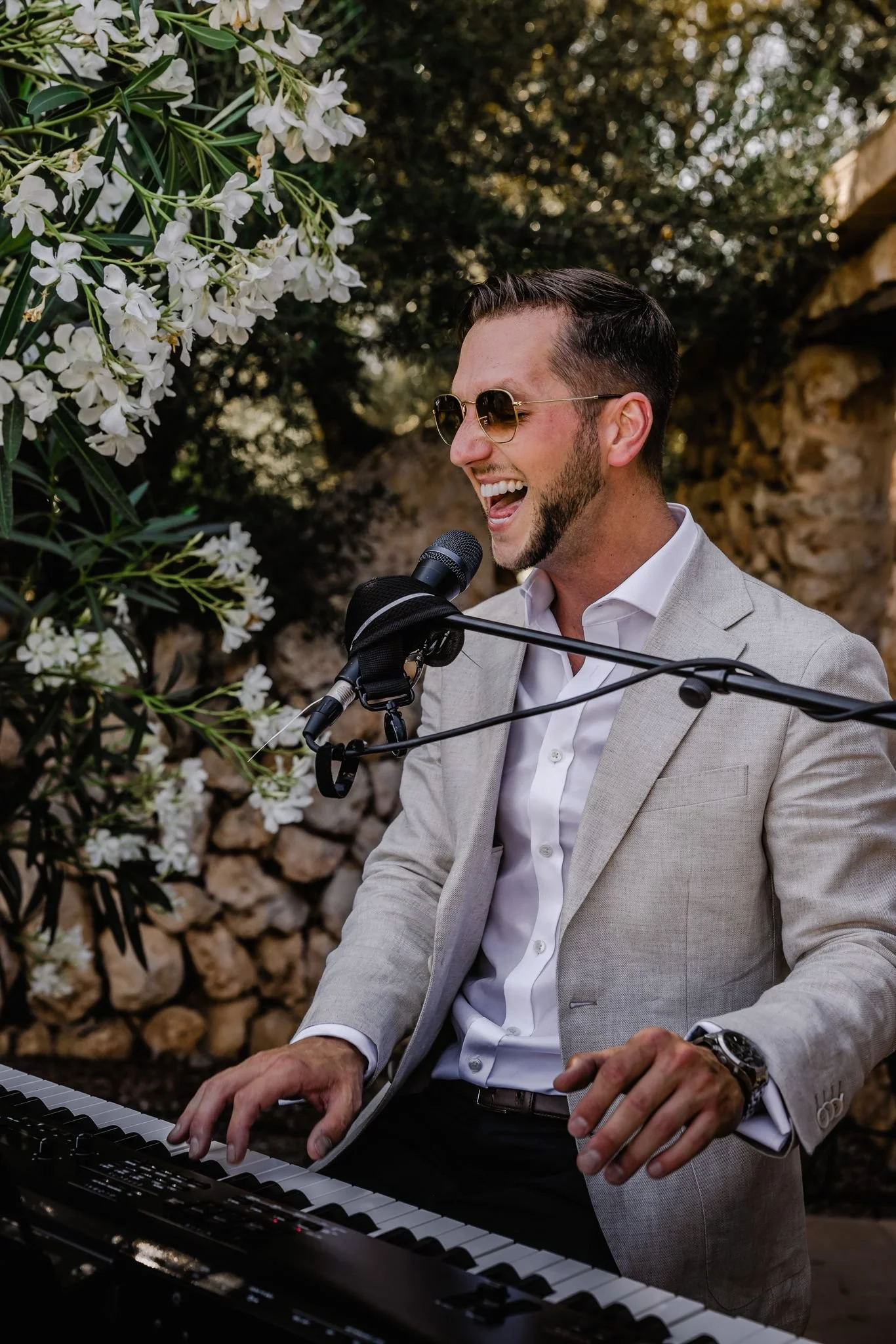 A man in a light gray suit and sunglasses plays a keyboard and sings into a microphone outdoors, smiling. There are white flowers and a stone wall in the background.