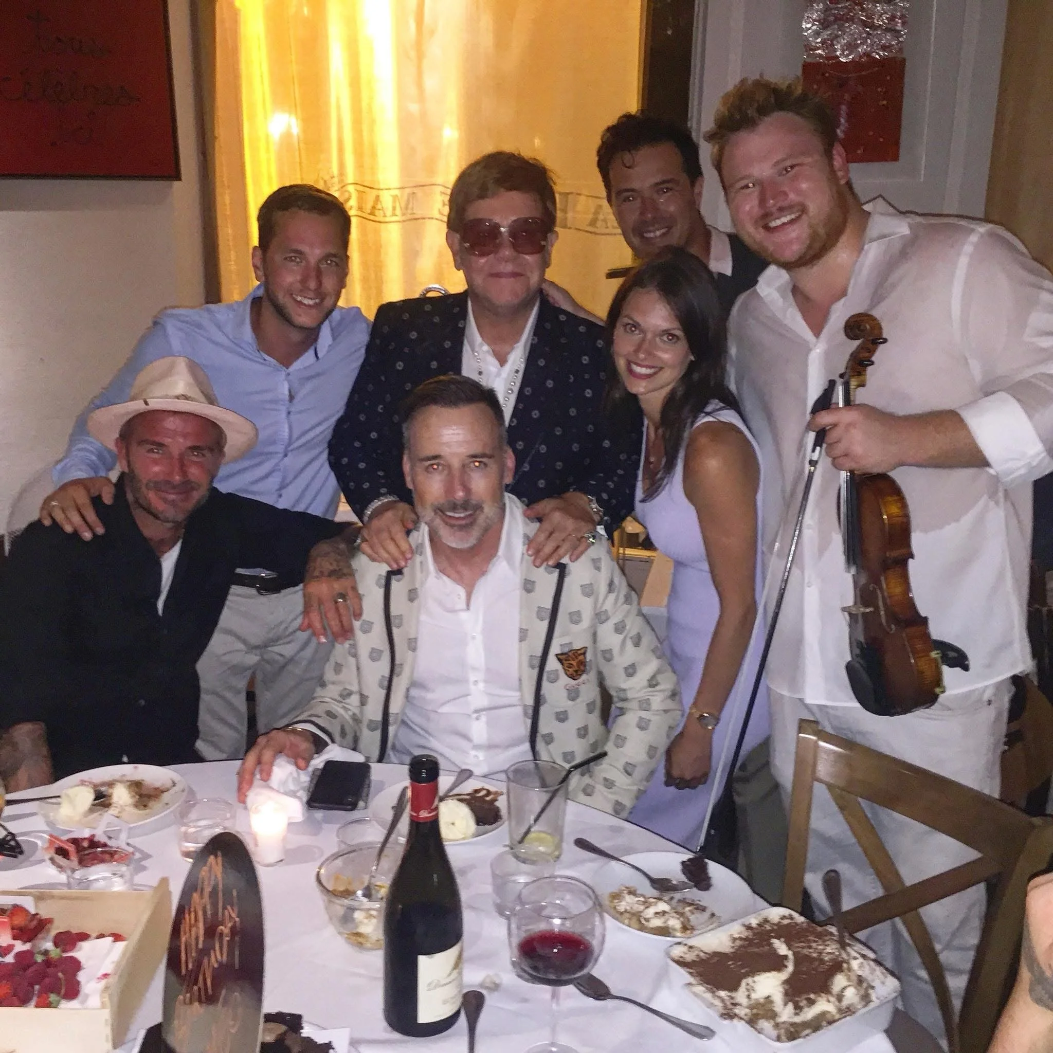 A group of eight people at a dinner party, smiling and posing for a photo. The table has desserts, wine, and a birthday cake.