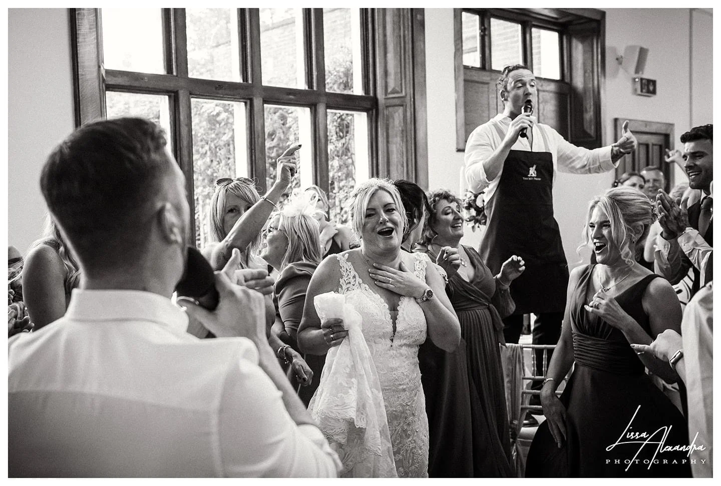 Black and white photo of a wedding reception. The bride is in the center, smiling and holding a cloth, with her hand over her chest. Guests around her are dancing, laughing, and pointing. A man in an apron is singing or speaking into a microphone on 