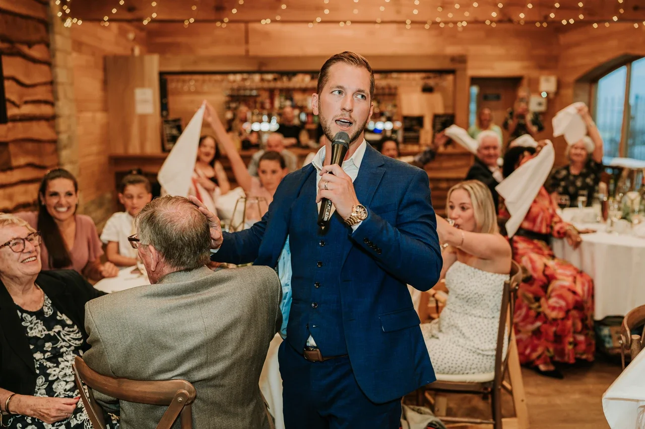 A man in a blue suit is speaking into a microphone at a wedding reception, touching the head of an elderly man seated at the table. Guests are smiling and cheering in a warmly decorated wooden banquet hall.