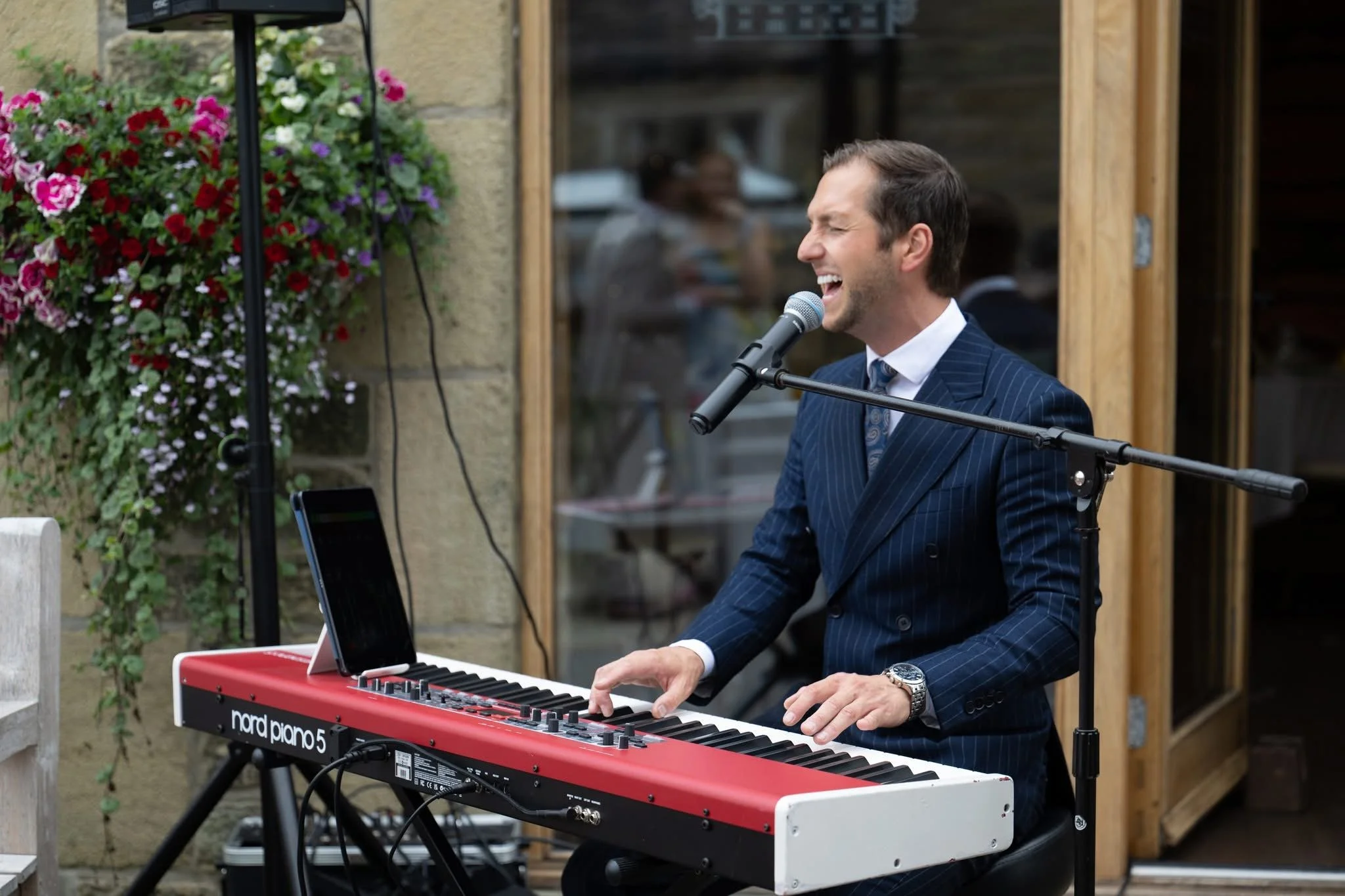 Man in a pinstripe suit playing a red and white Nord Piano keyboard and singing into a microphone outdoors.