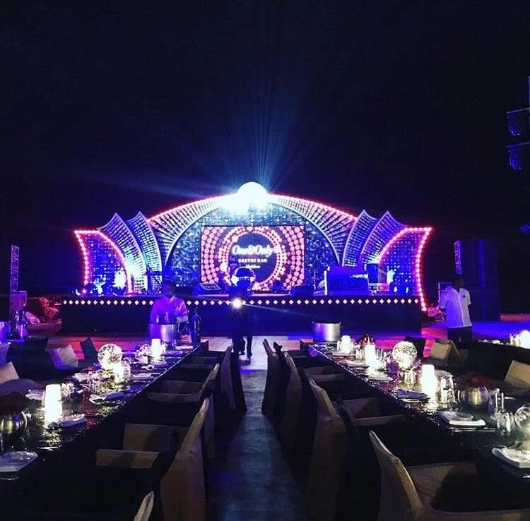 A nighttime event with a stage decorated with bright, colorful lights and a large circular screen displaying text. The stage has artistic arches and a central bright light. In the foreground, there are tables with chairs, candles, and tableware set for guests.