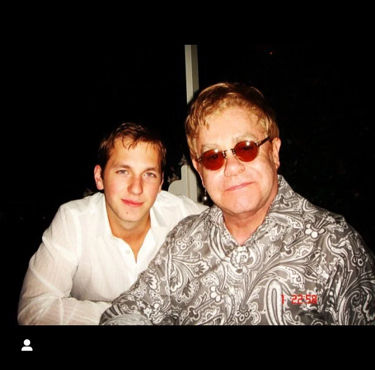 Two men sitting together, one wearing a white shirt and the other wearing a patterned shirt and sunglasses, during nighttime.