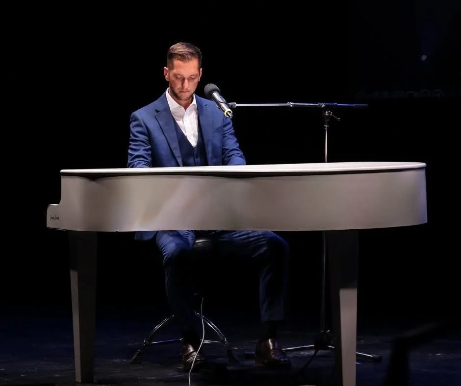 A man in a blue suit playing a white grand piano on stage with a microphone positioned above him.