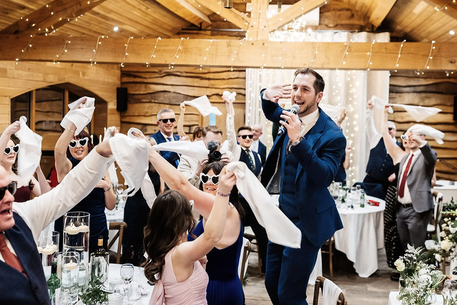 Man in a blue suit singing and dancing with guests at a wedding reception in a rustic venue decorated with string lights.