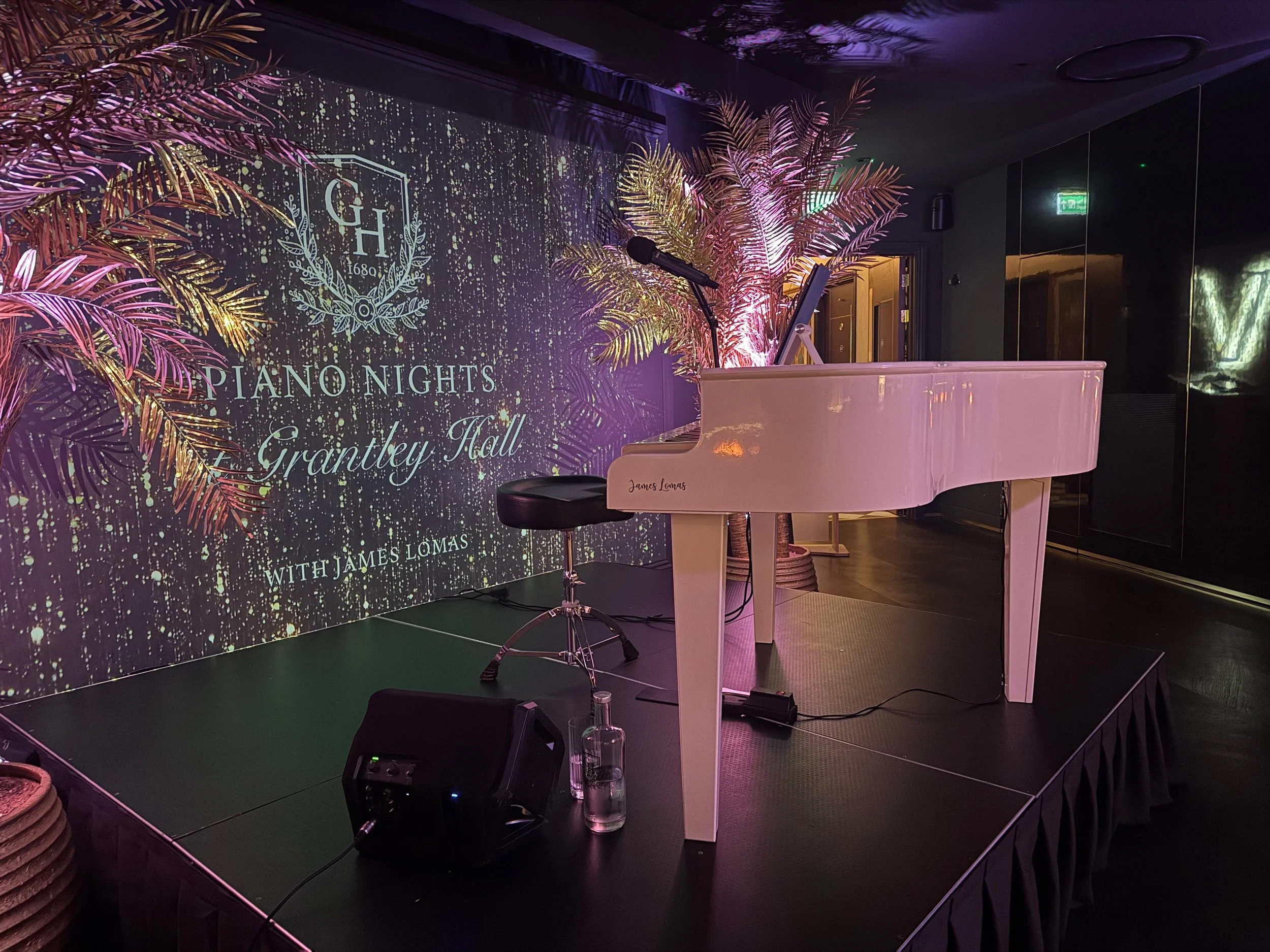 White grand piano on a stage with a microphone, a black stool, and a speaker. Behind the piano, a large illuminated graphic screen displays a logo for Piano Nights at Grantley Hall with purple and gold lighting. Decorative pink and gold palm trees are on the stage, creating an elegant setting for a musical performance.