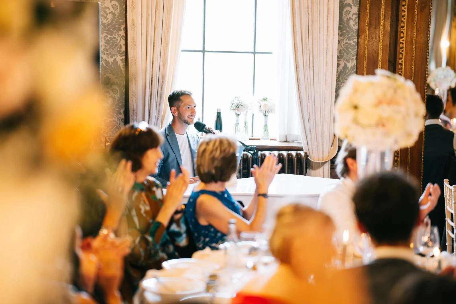A man giving a speech at a formal event with audience members applauding, set in an elegantly decorated room with large windows and floral arrangements.