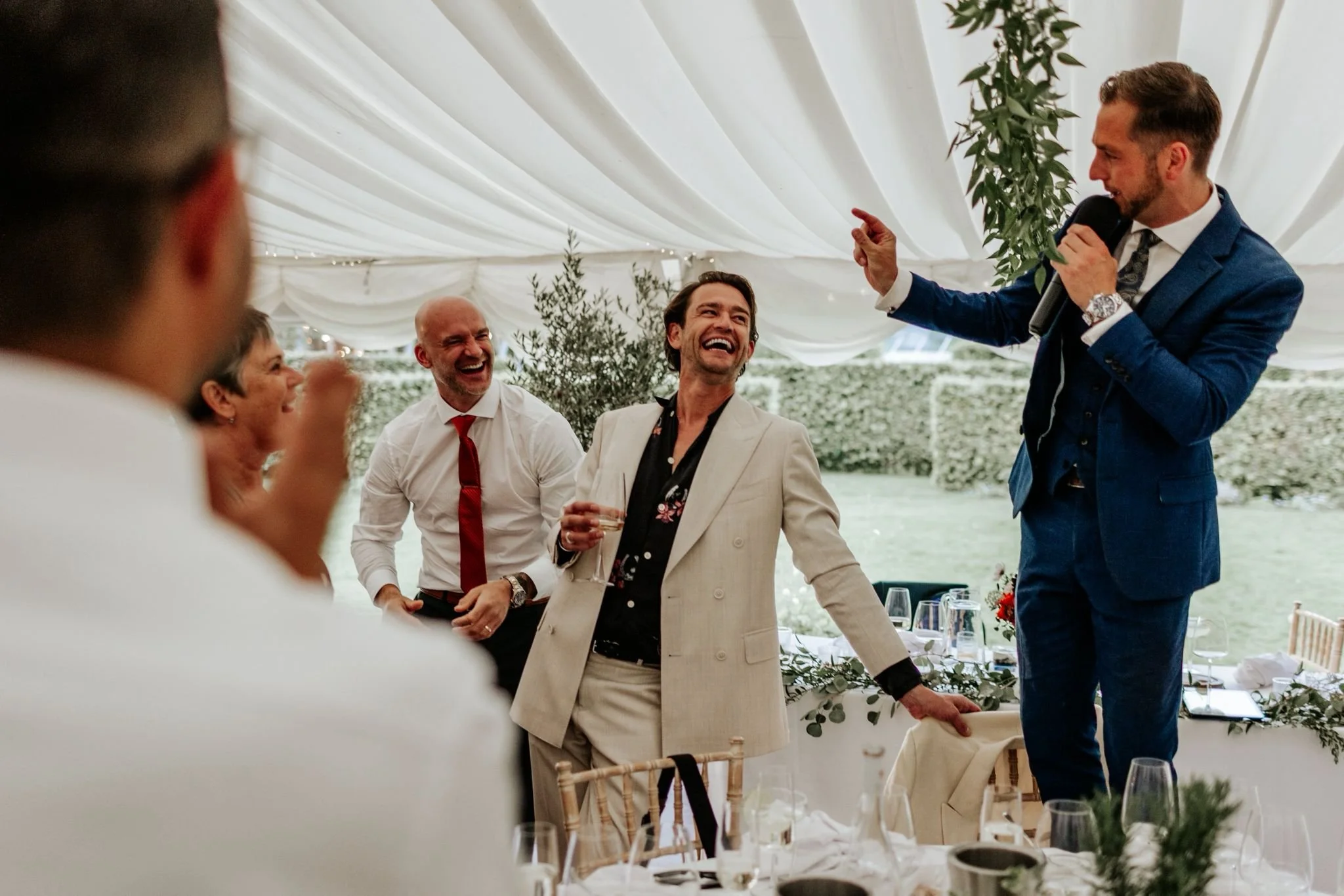 People at a wedding reception/community celebration inside a decorated tent, with one man giving a speech or toast, while others laugh and enjoy the moment.