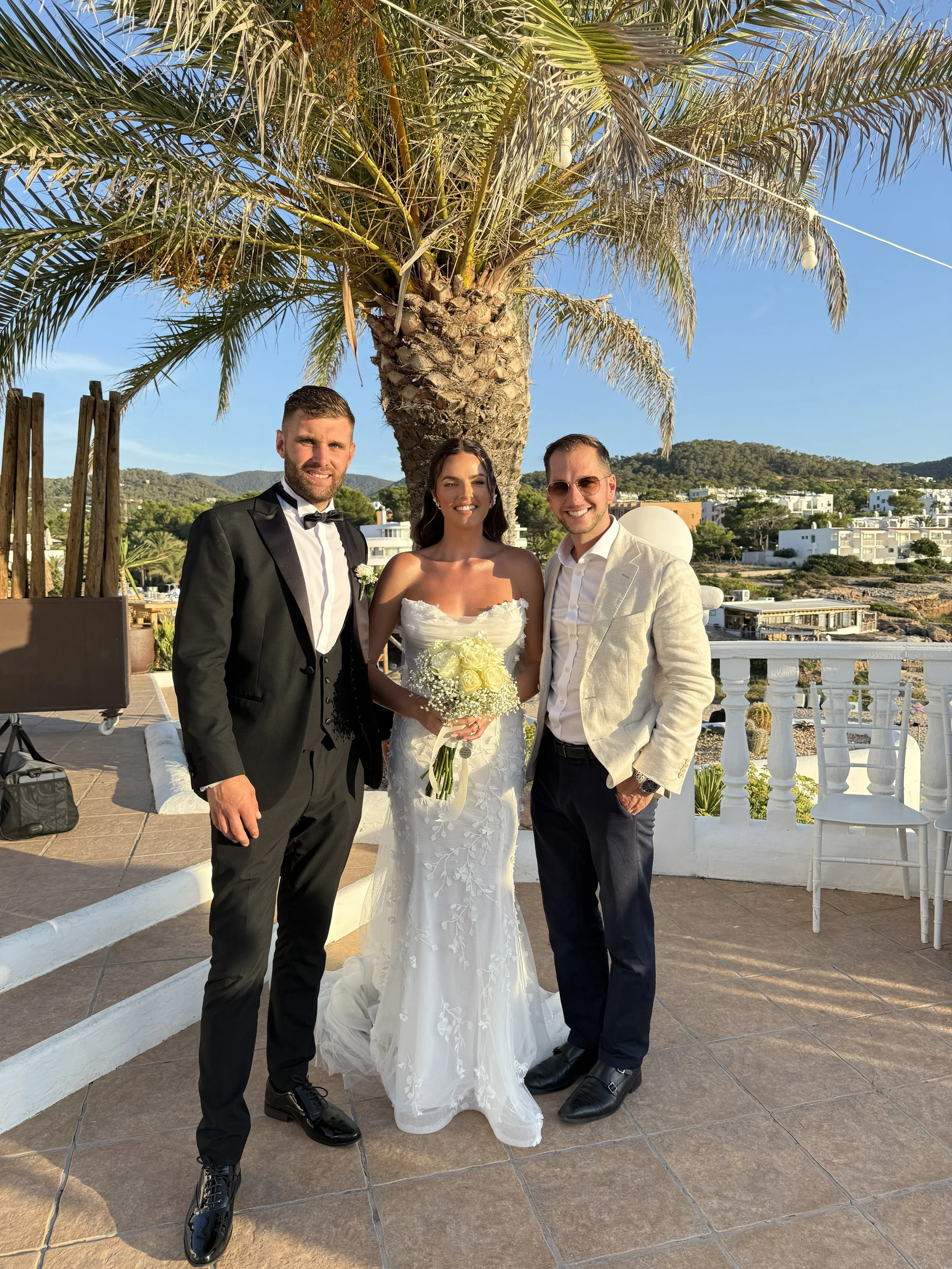 Three people smiling at a wedding, standing outdoors near a palm tree, with a scenic town view in the background. The bride holds a bouquet of white flowers, with a white wedding dress, and the two men wear suits.