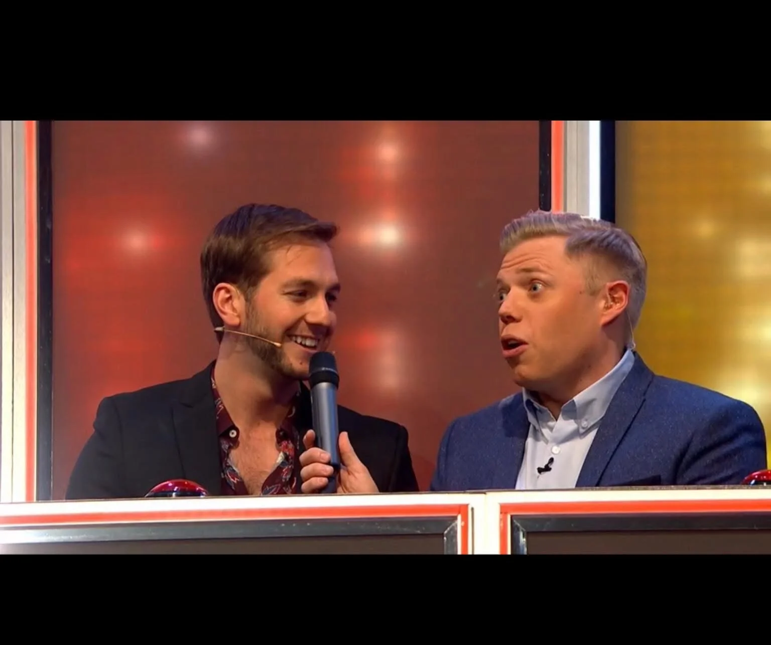 Two men on a game show stage, one holding a microphone and wearing a headset, the other speaking with a surprised expression, with a red and gold illuminated background.