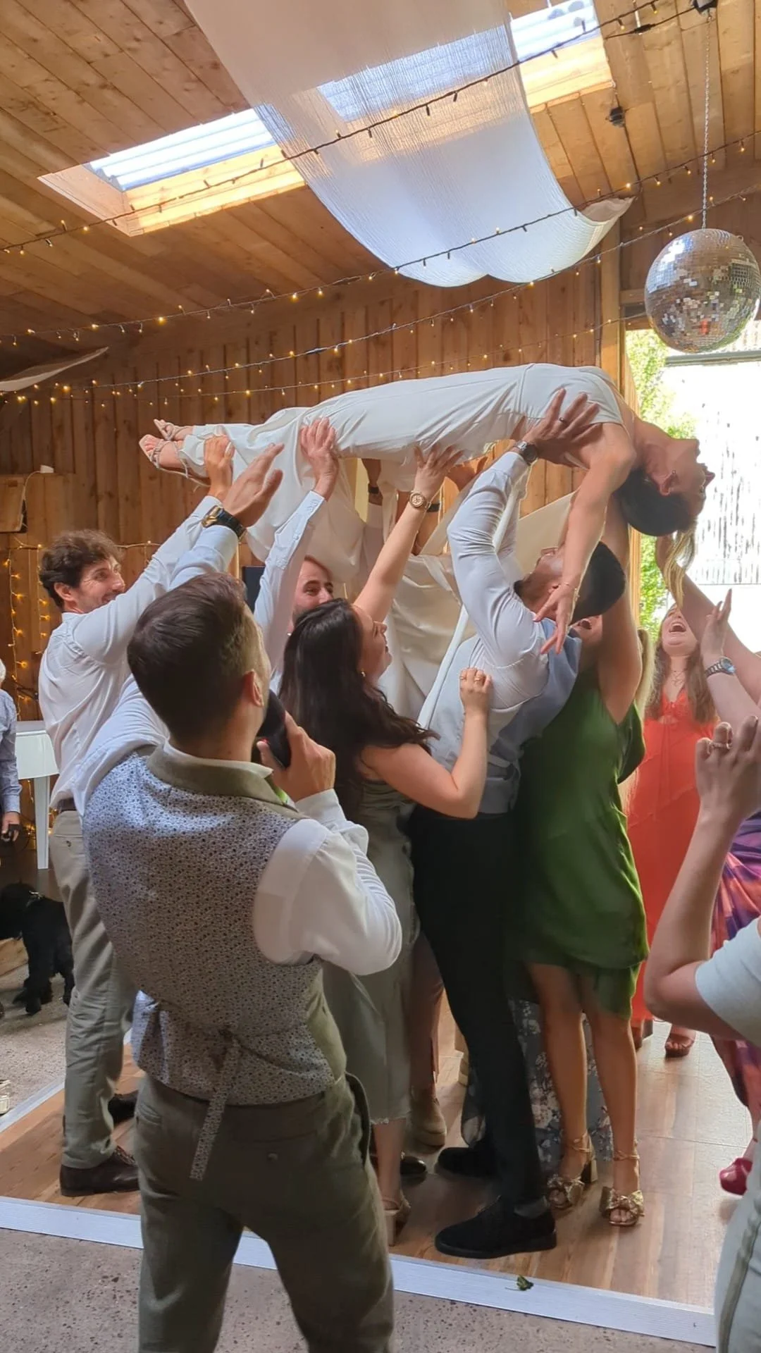 Group of people at a celebration lifting a woman in a white dress in the air, with a decorated wooden ceiling, string lights, and a disco ball in the background.