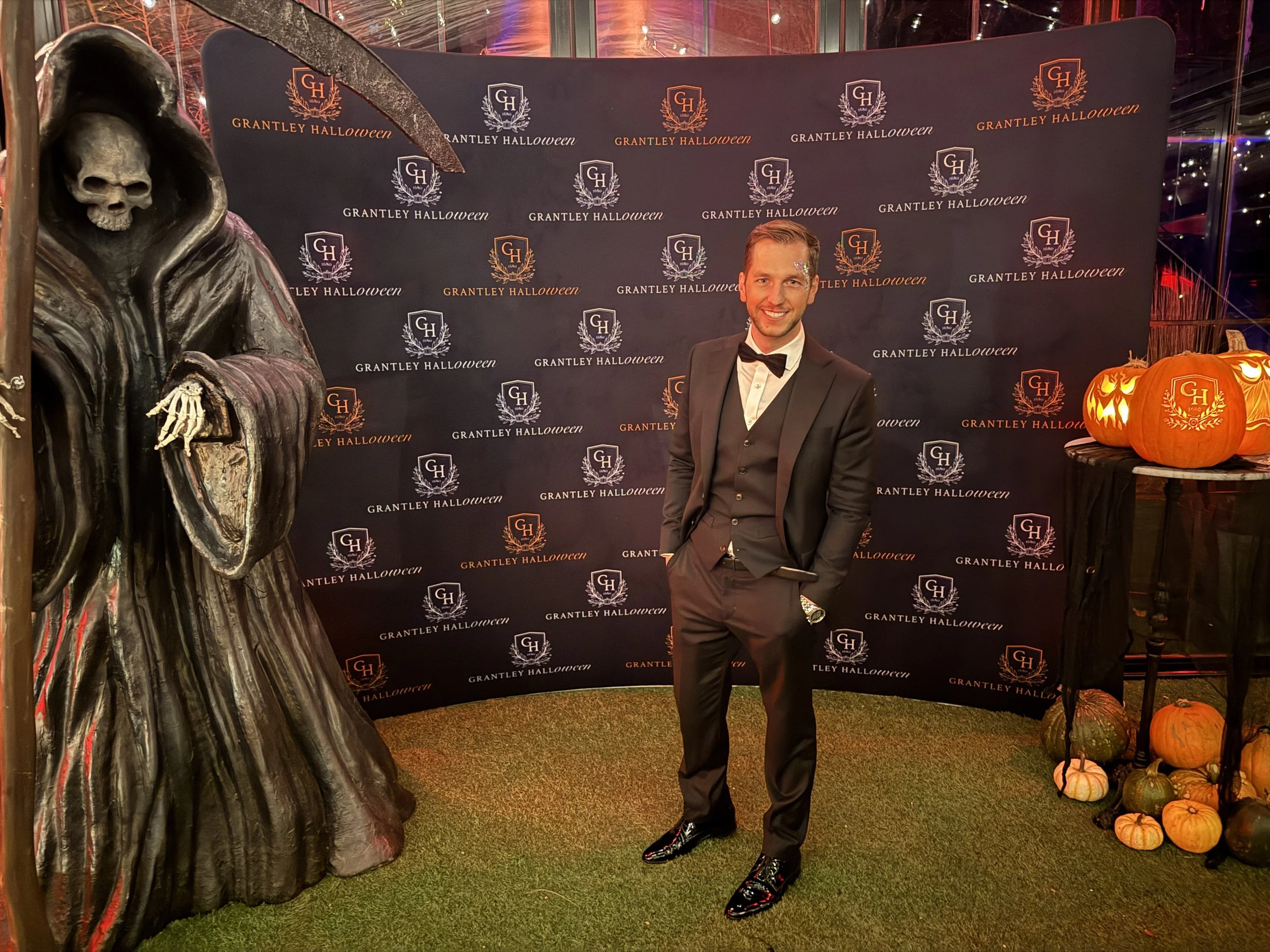 A man in a black tuxedo with a bow tie, standing in front of a backdrop with 'Grantley Halloween' logos, smiling, with Halloween-themed decorations including a large skeleton figure and orange pumpkins, some decorated with 'G H' logos, at a Halloween