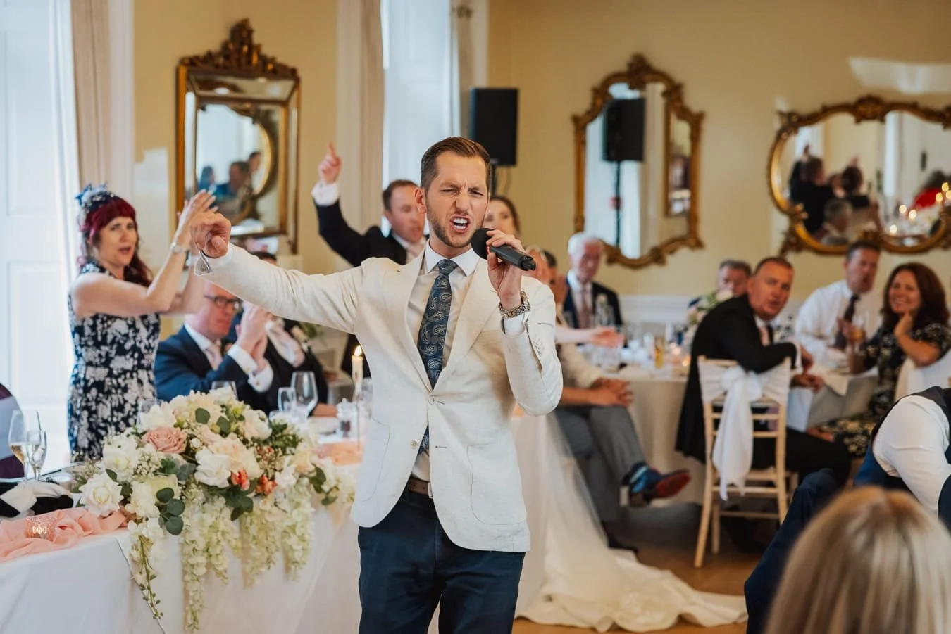 Man singing passionately at a wedding reception, surrounded by seated guests in a decorated banquet hall with mirrors.