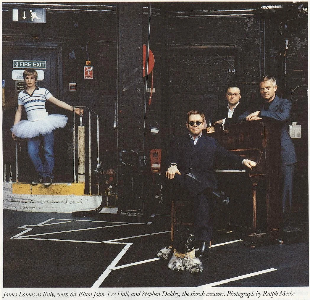 A young girl in a tutu and striped shirt stands at a door with a fire exit sign, holding a leash. Three men dressed in dark suits and sunglasses sit and stand around a vintage piano in a dark, industrial space. The caption indicates they are performe