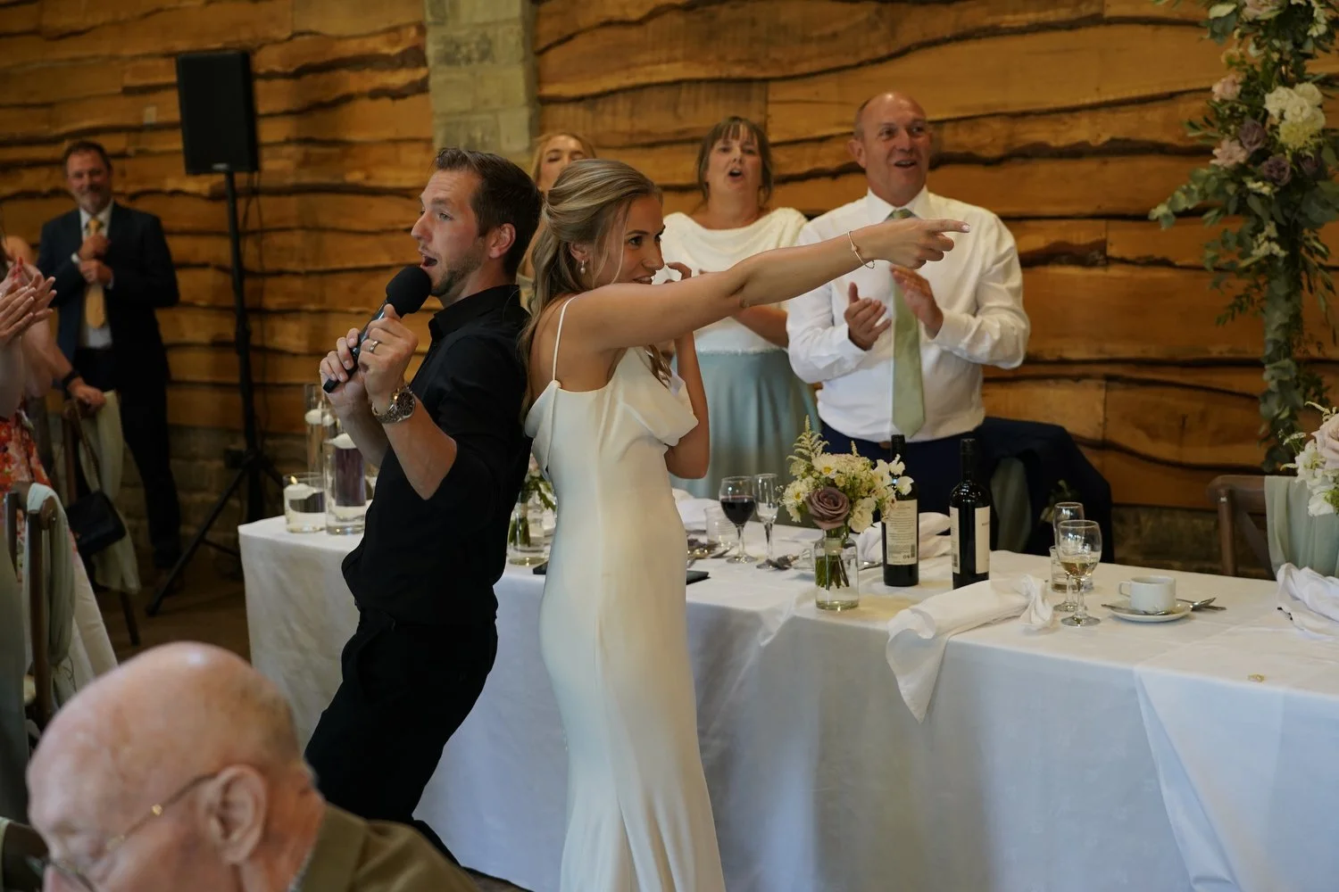 A wedding reception with a bride and groom singing and dancing, guests applauding and watching in a rustic wooden venue.