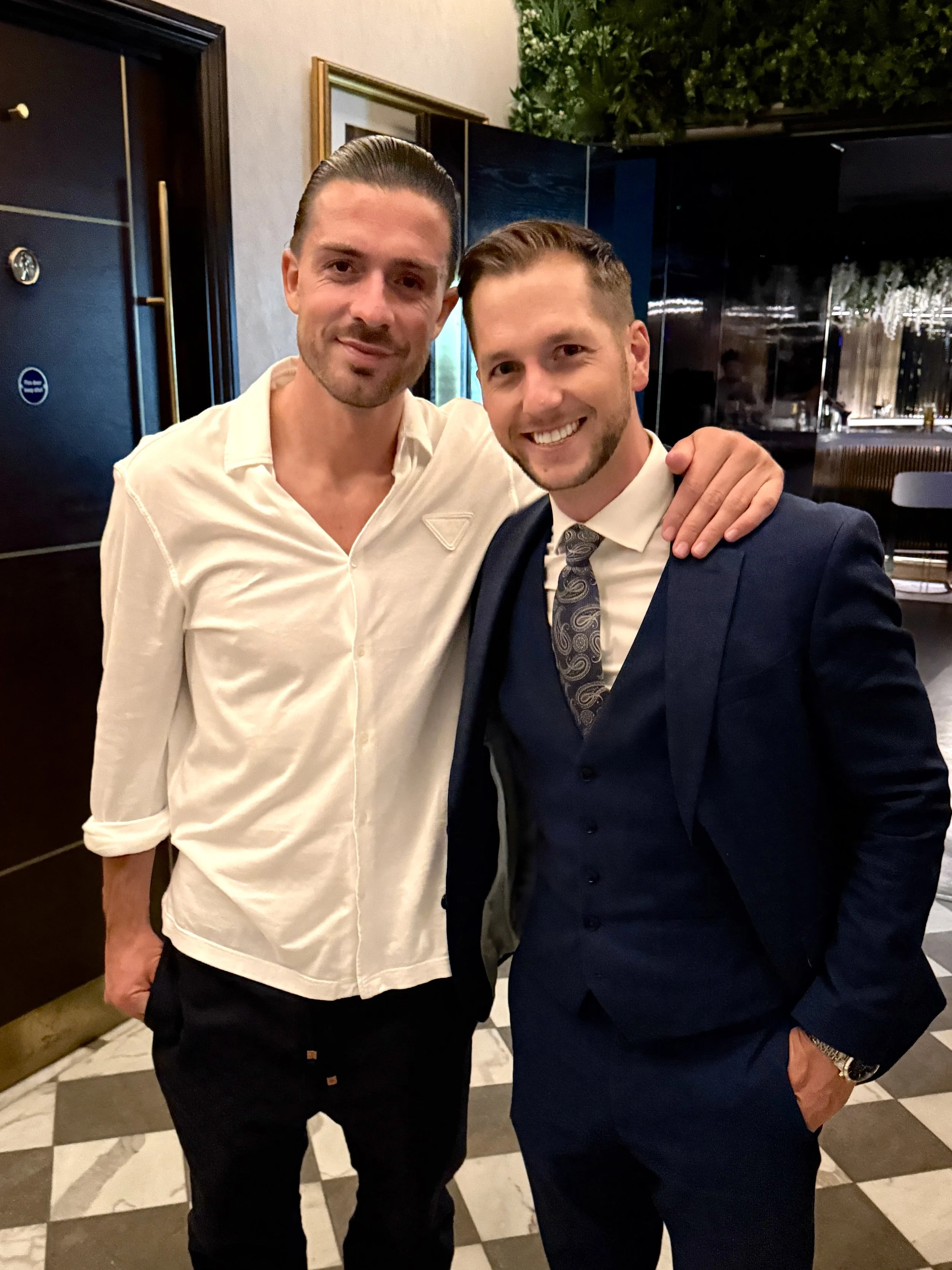 Two men standing close together indoors, smiling, one in a white shirt and the other in a navy suit with a patterned tie.