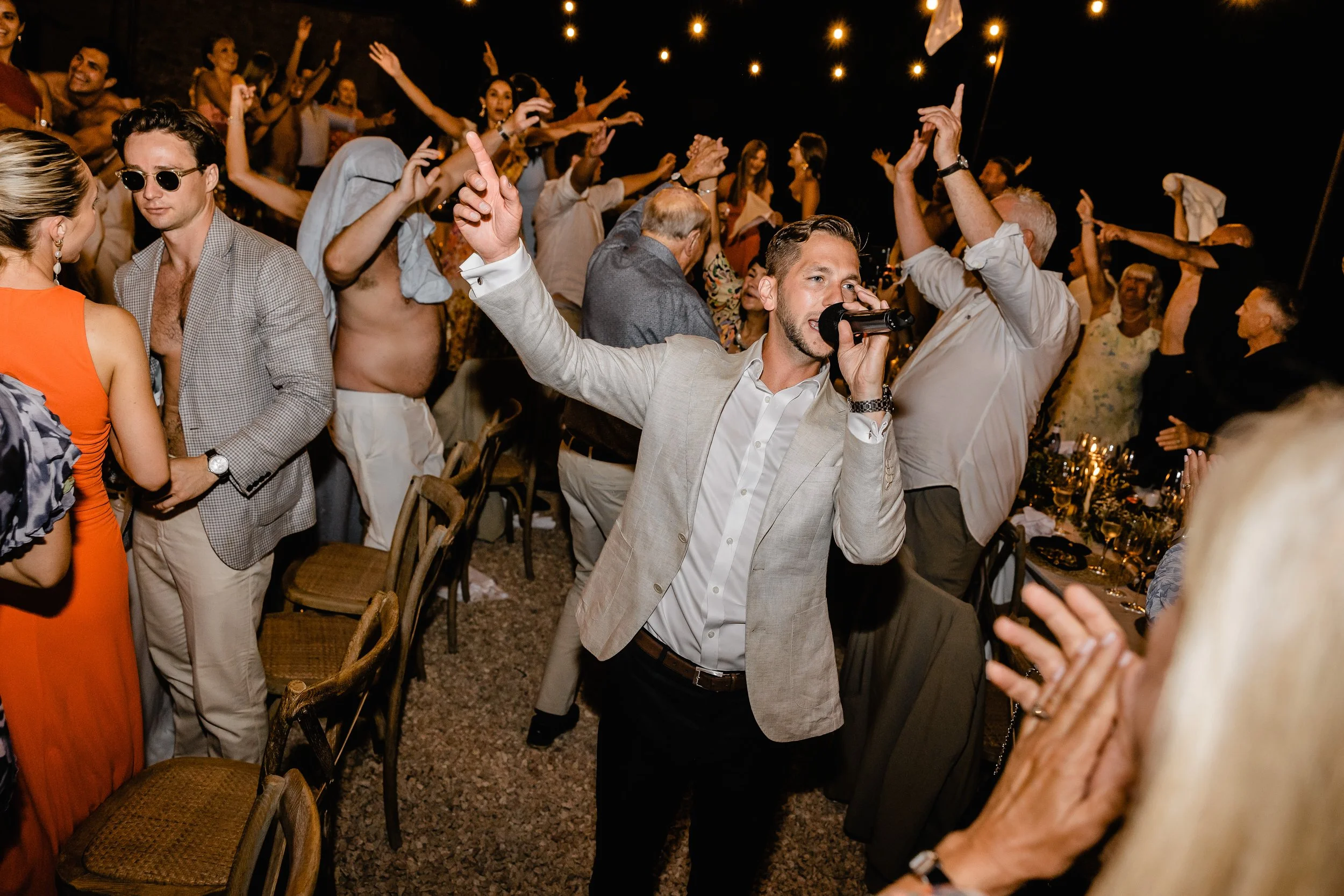 Man singing into a microphone at a lively indoor celebration with dancing and people clapping and raising their hands under hanging string lights.