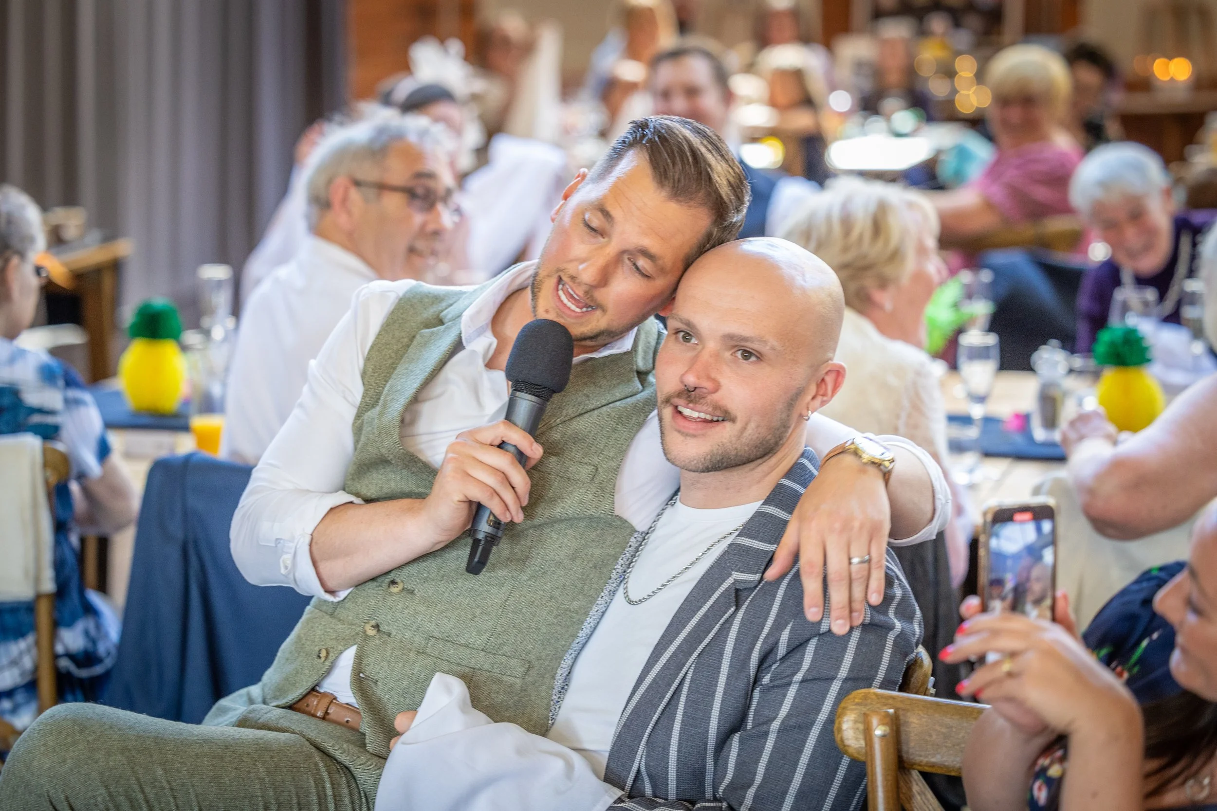 Two men at a social gathering, one holding a microphone, with people seated around in the background.