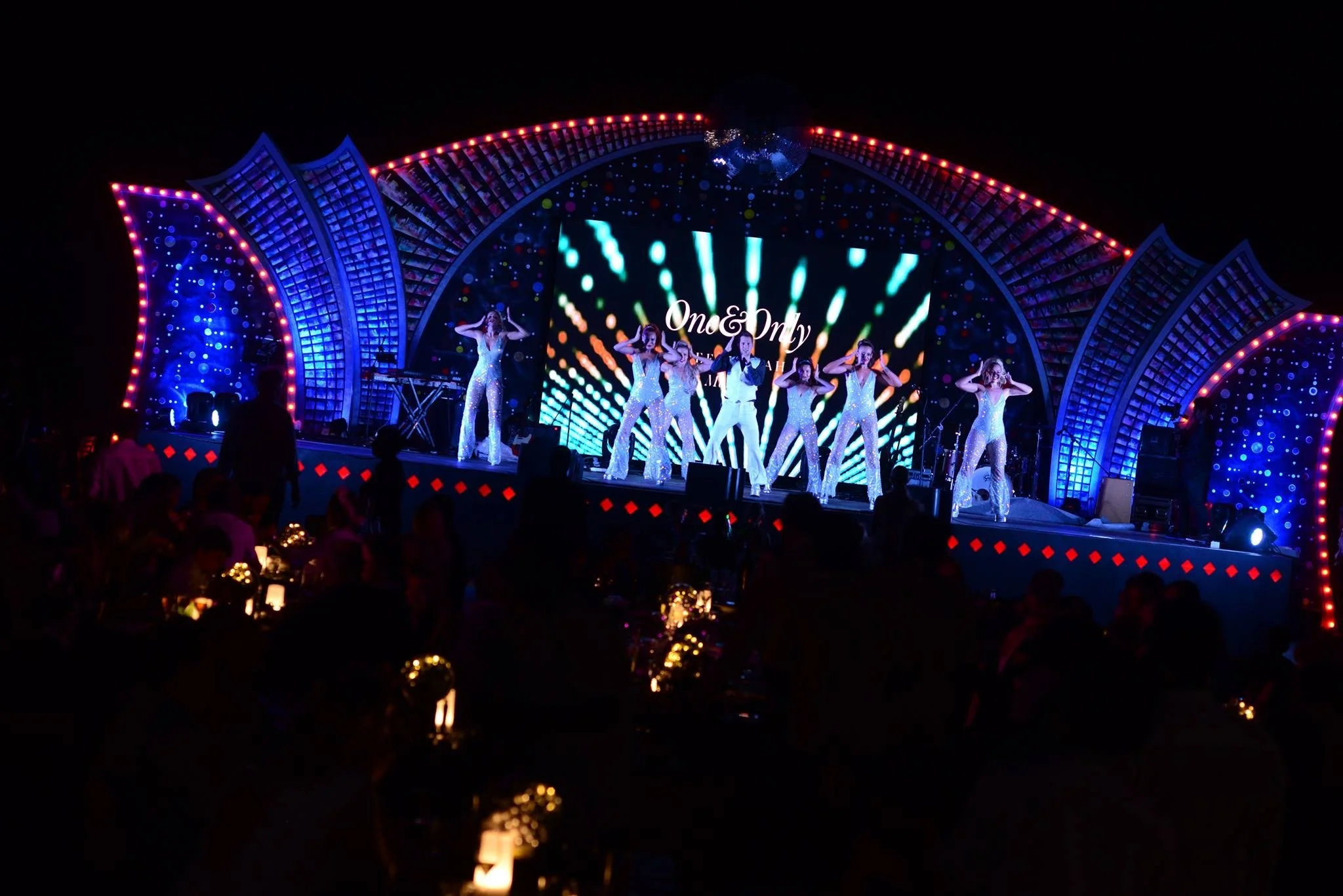A live stage performance at night with dancers in silver costumes, with colorful LED screens and lighting, and an audience seated at tables with candles.