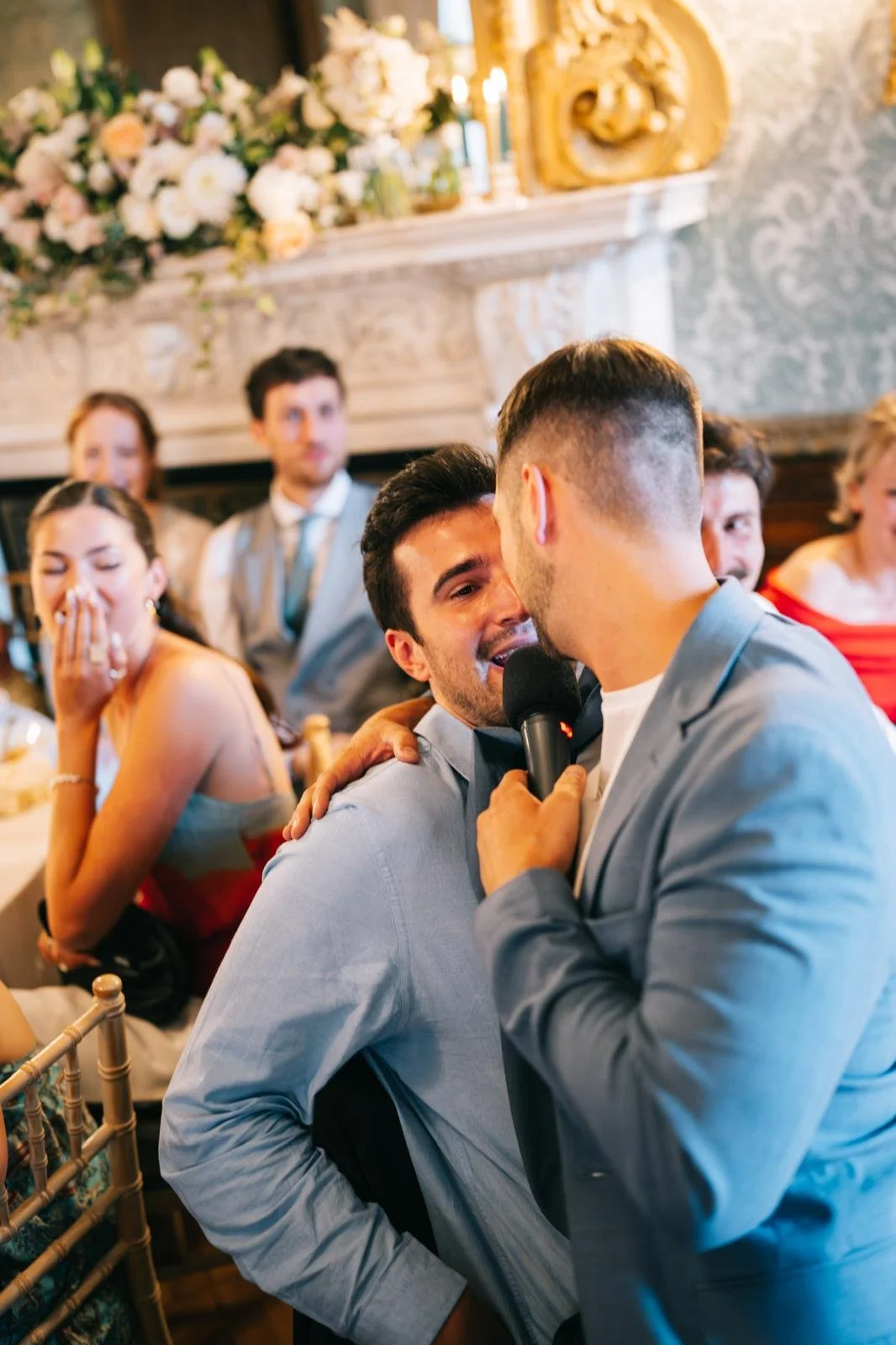 A man in a gray blazer is holding a microphone close to another man, who appears to be emotional or crying, during a speech or toast at a formal gathering. Several guests, including women and men, are seated around a decorated table, some with expres