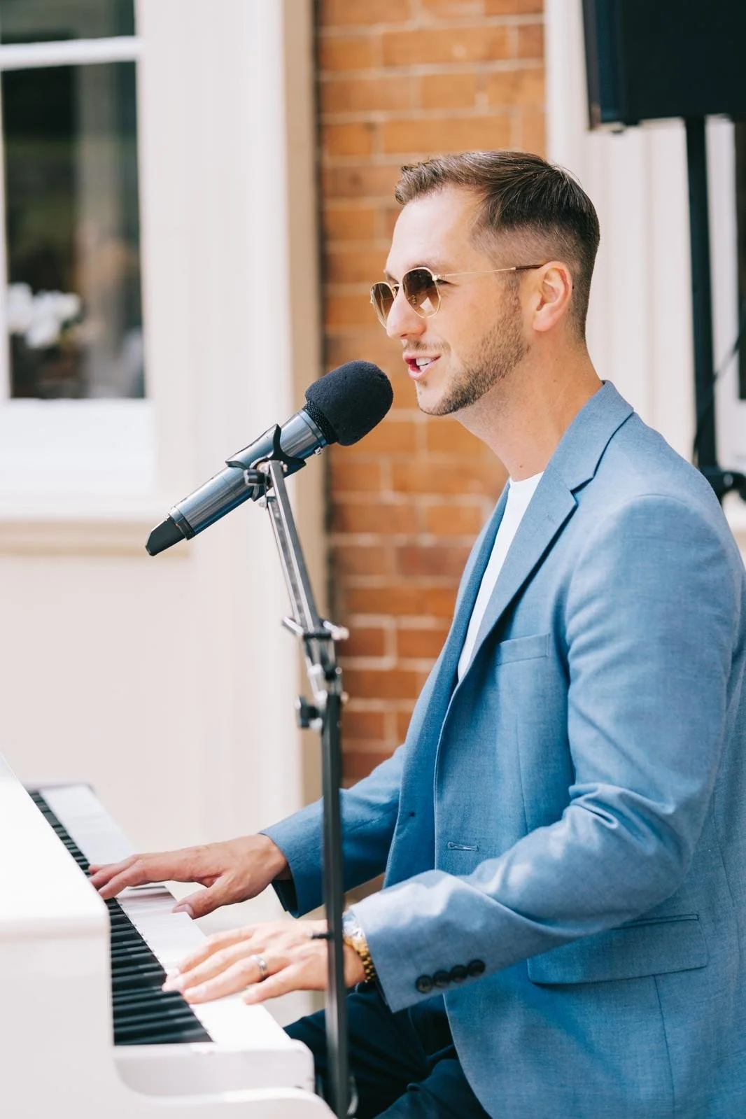 A man wearing sunglasses in a blue blazer playing a white piano and singing into a microphone outdoors.