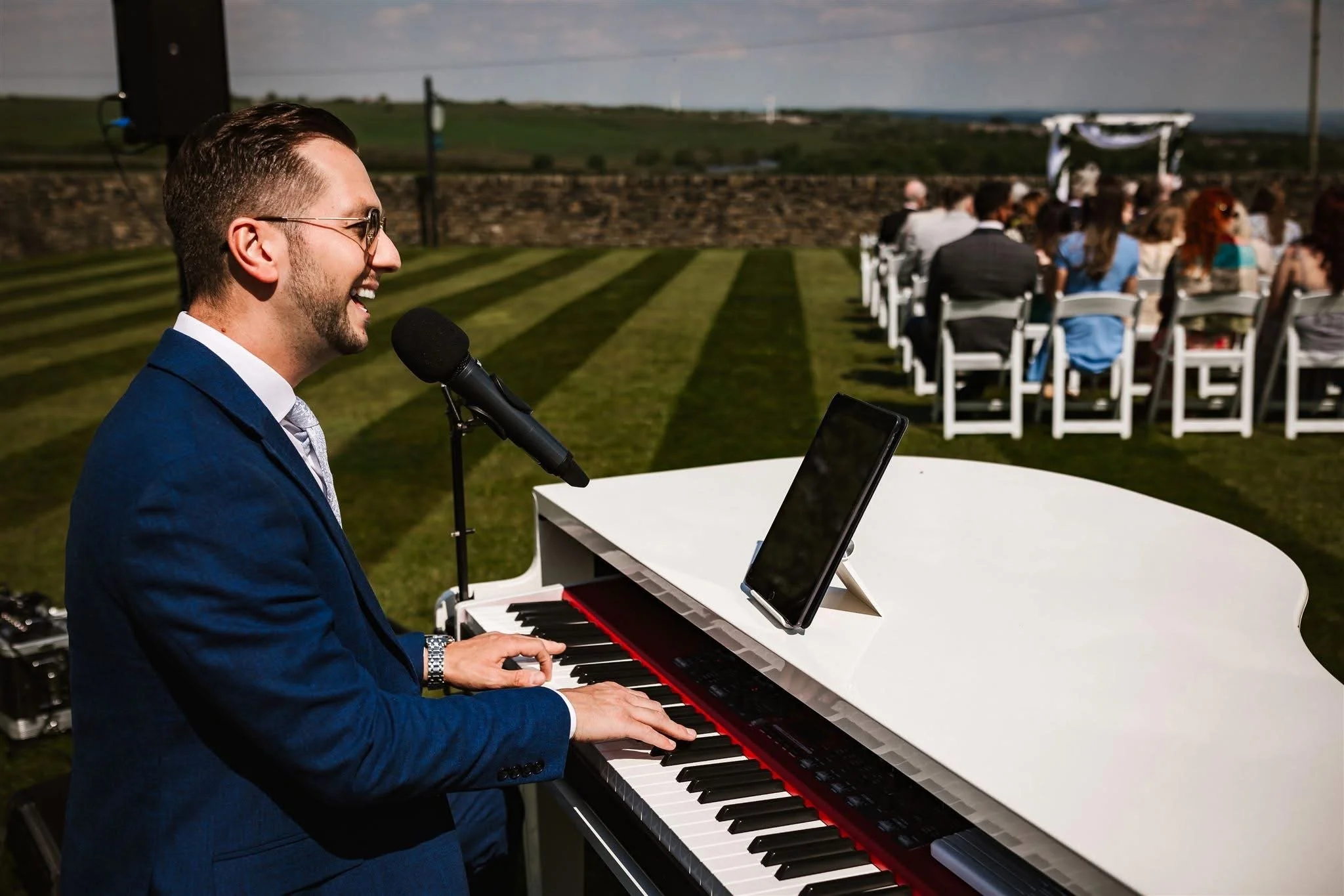 A man in a blue suit playing a white grand piano outdoors during a wedding ceremony, with guests seated in rows behind him.