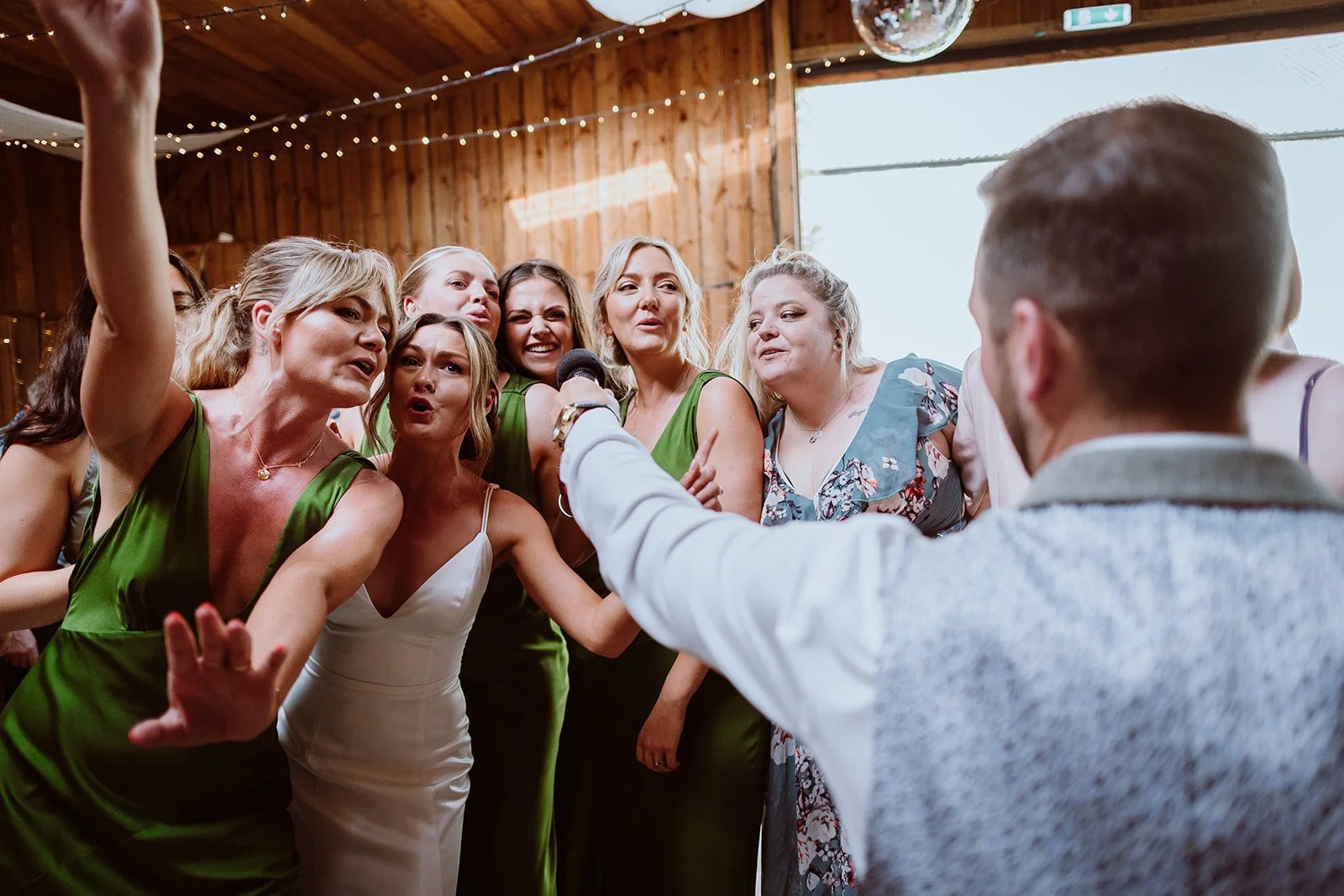 A group of women singing and dancing together at a celebration, with a man holding a microphone facing them in a wooden room decorated with string lights.