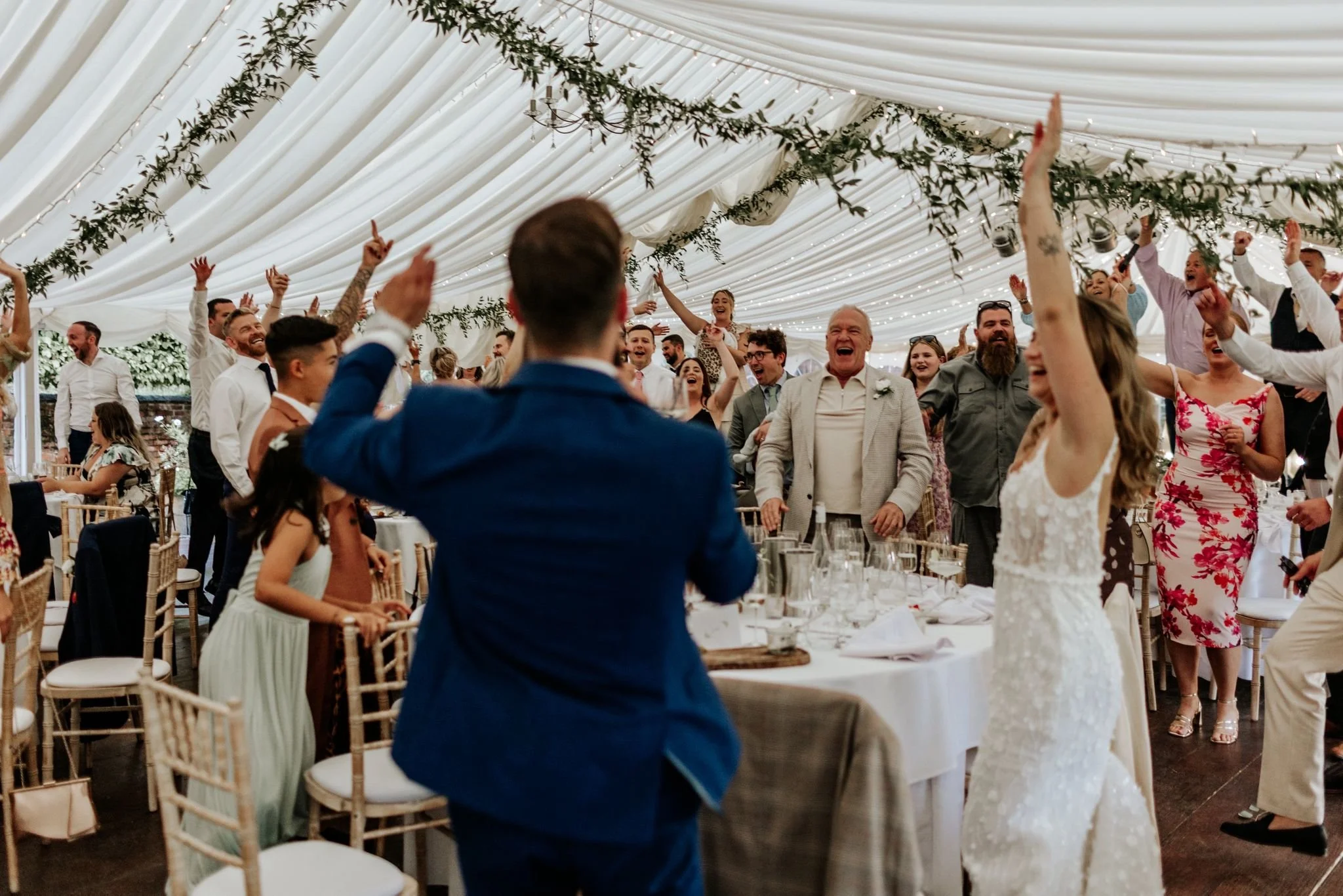 Celebrating wedding with guests dancing and cheering under a decorated white tent.