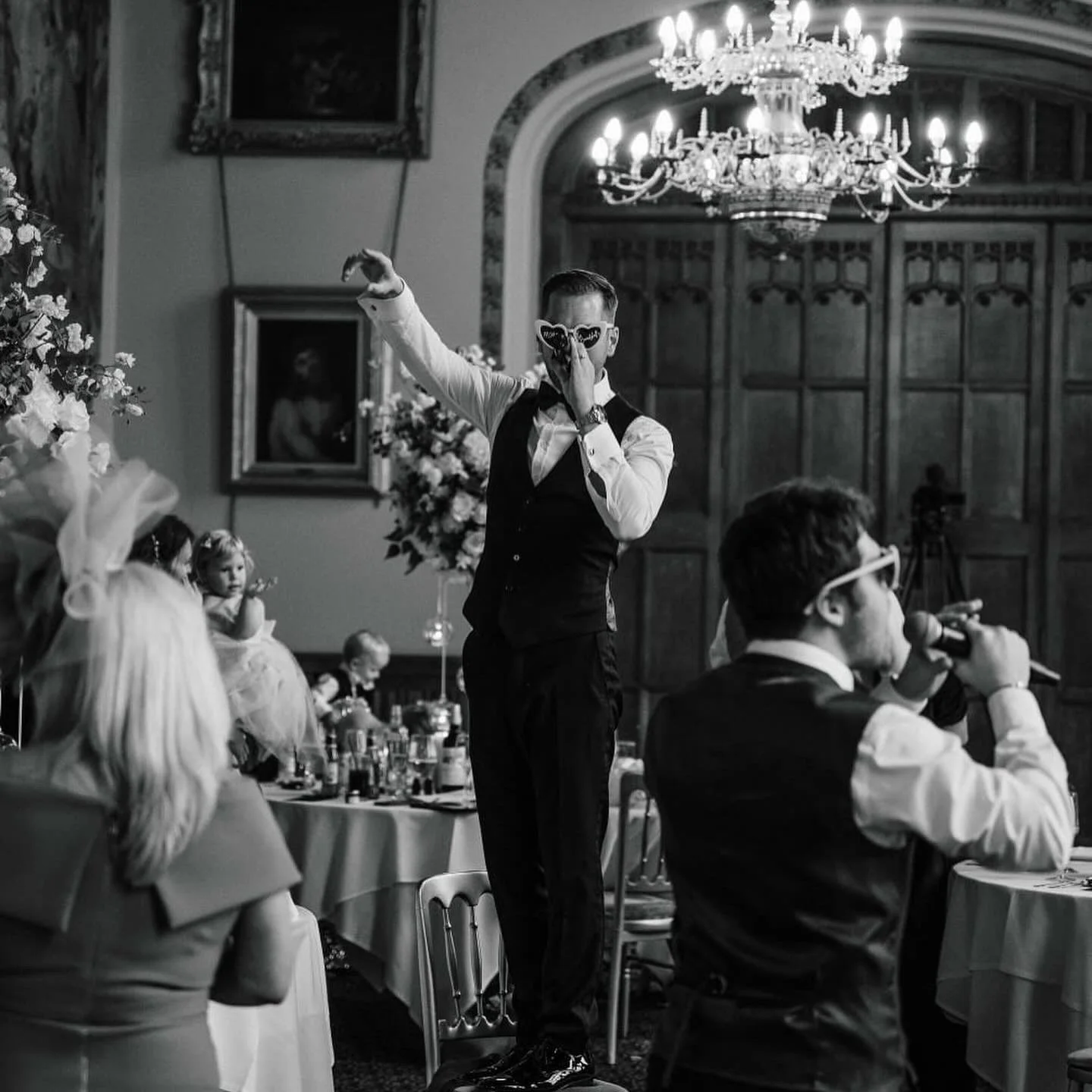 A man in formal attire and sunglasses stands on a chair, pointing upwards and holding his nose, at an indoor event with other people seated at tables, including children in fancy dresses, with a chandelier and framed paintings in the background.