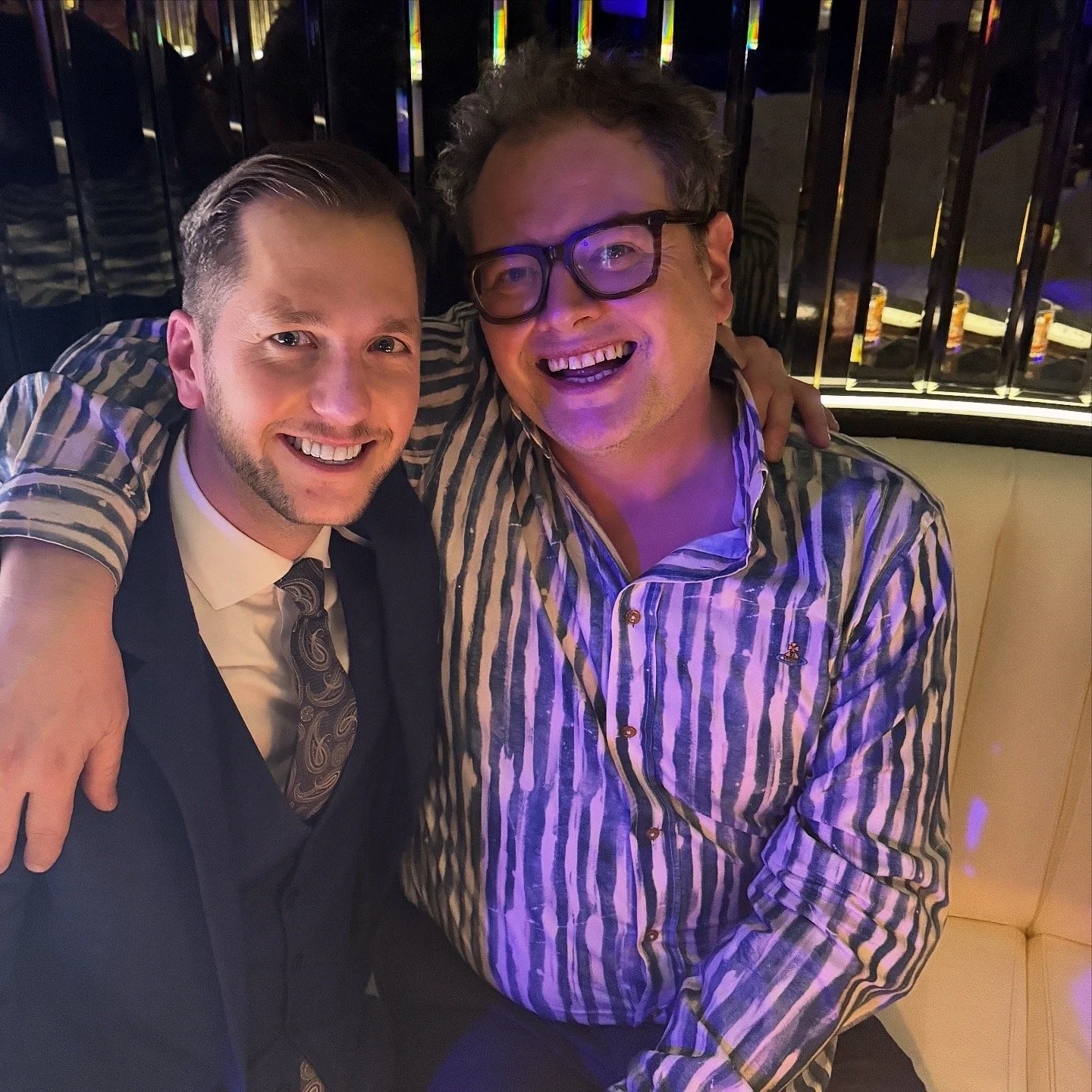 Two men smiling and sitting close together in a festive setting, one with arm around the other, with colorful lighting and a reflective backdrop.