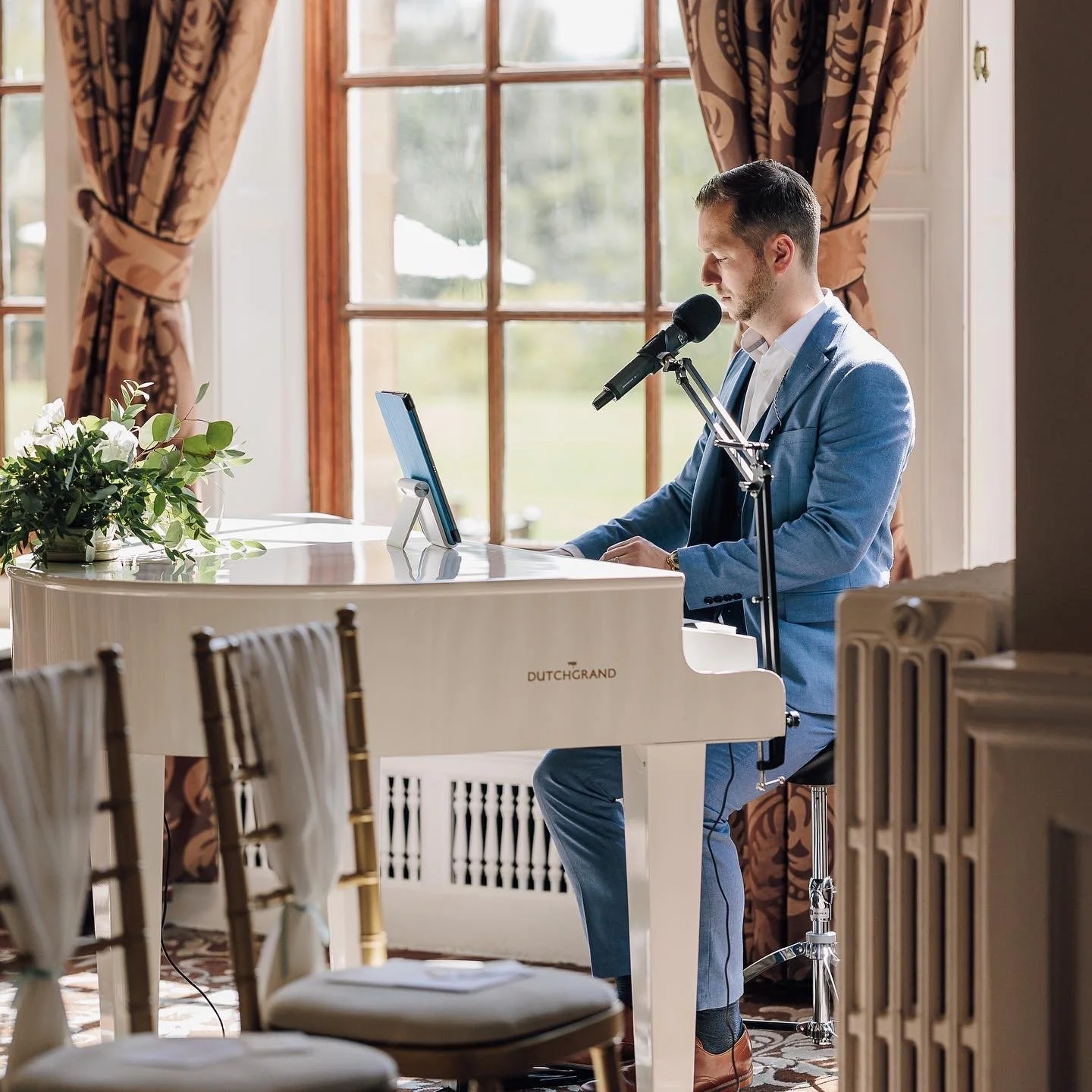 A man in a blue suit playing a white grand piano in a room with large windows and brown patterned curtains.