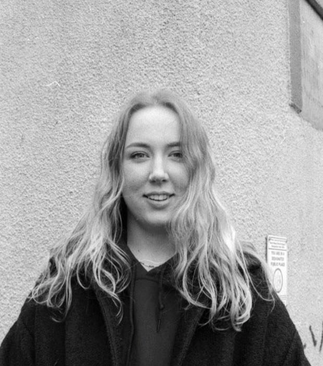 A woman with wavy hair wearing a dark hoodie and jacket standing against a textured wall.