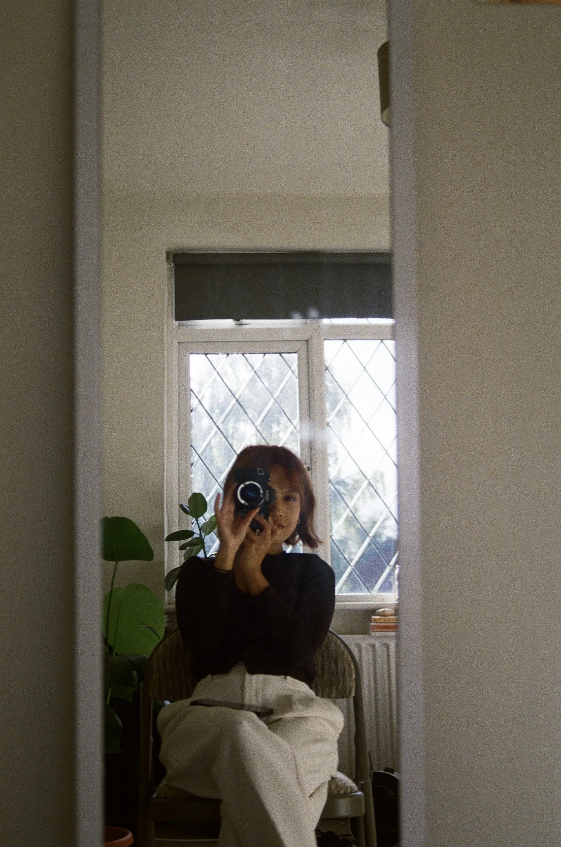 A woman with short hair takes a mirror selfie with a camera, sitting on a chair in a room with large diamond-pane windows and a houseplant.