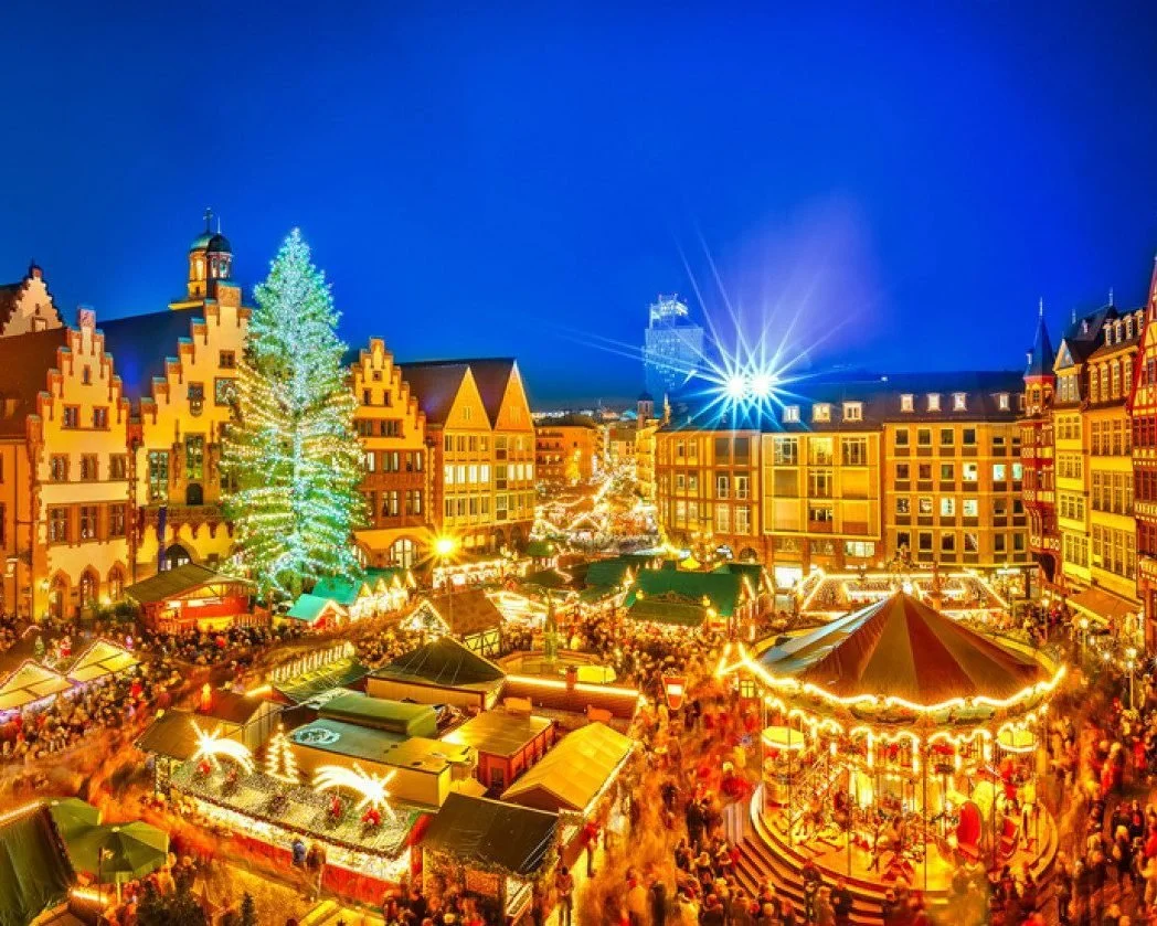 A lively Christmas market at night with festive lights, a large decorated Christmas tree, an ornate carousel, and historic European-style buildings in the background.