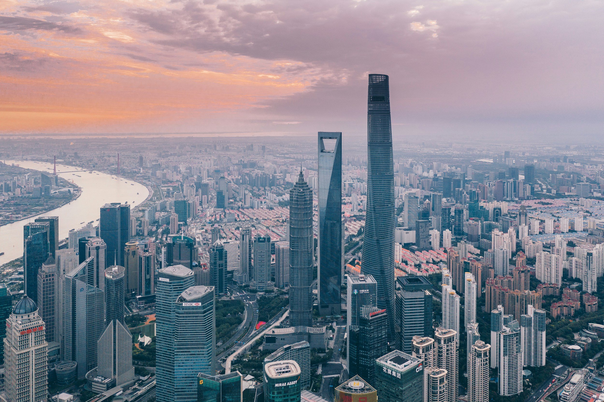 Aerial view of a city skyline at sunset with tall modern skyscrapers, a river, and a colorful sky with clouds.