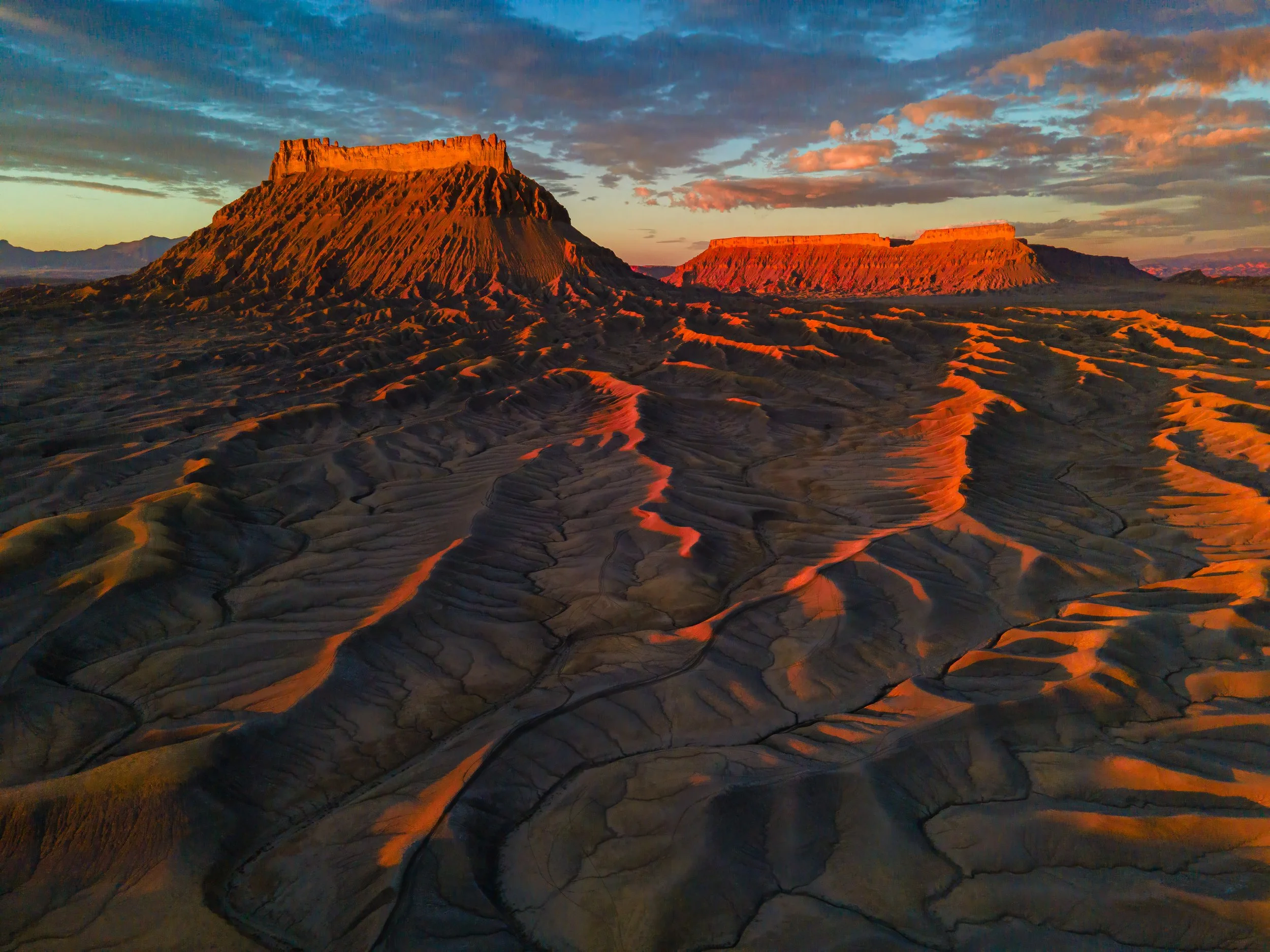 A desert landscape at sunset with two large flat-topped mesas and intricate sand formations in the foreground.