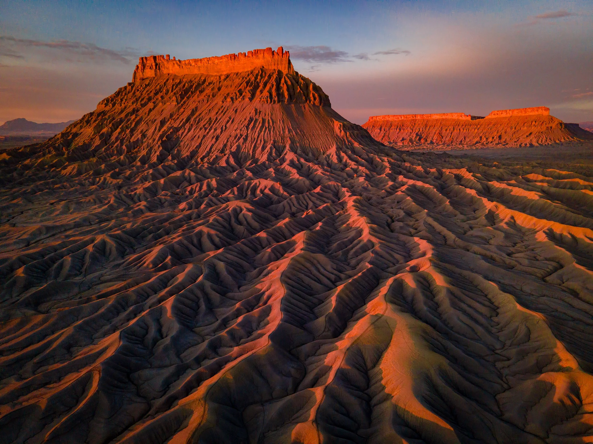 Scenic view of layered mesas and buttes in a desert at sunset, with the main mesa in the foreground illuminated by the setting sun.