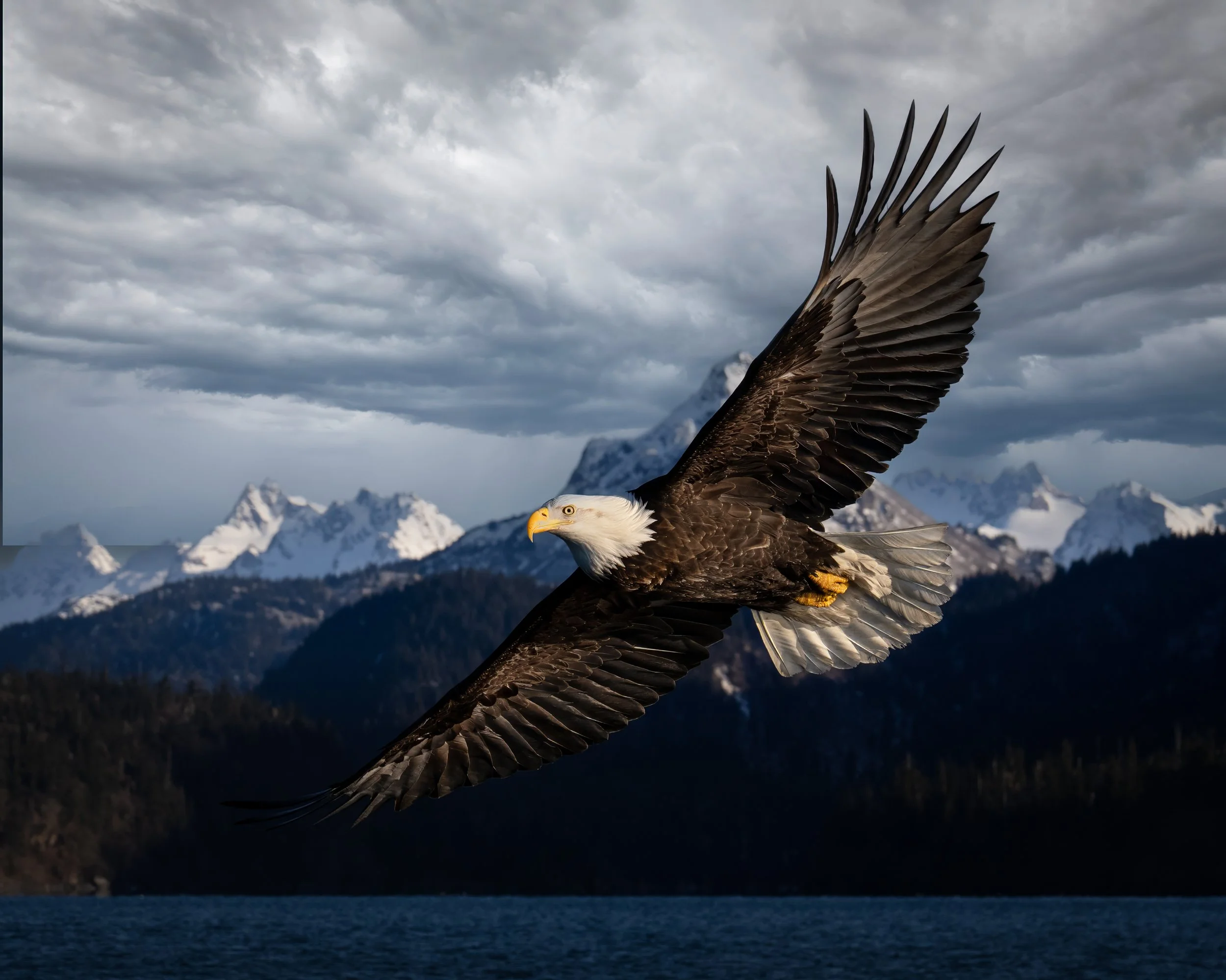 A bald eagle flying over a body of water with snow-capped mountains and cloudy sky in the background.