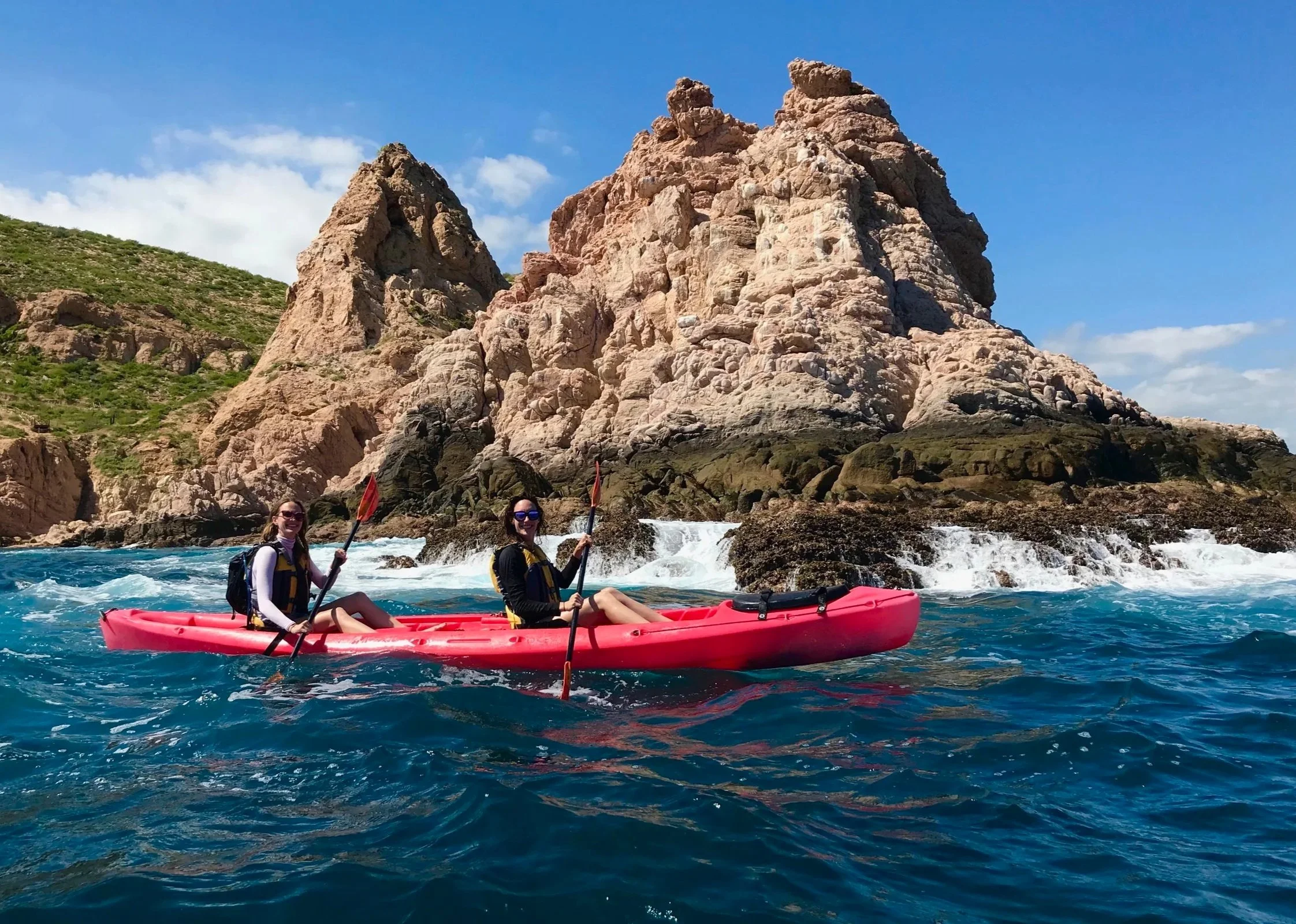 Two women in a red kayak paddling in the ocean near rocky cliffs under a blue sky on a Studio One Yoga Retreat