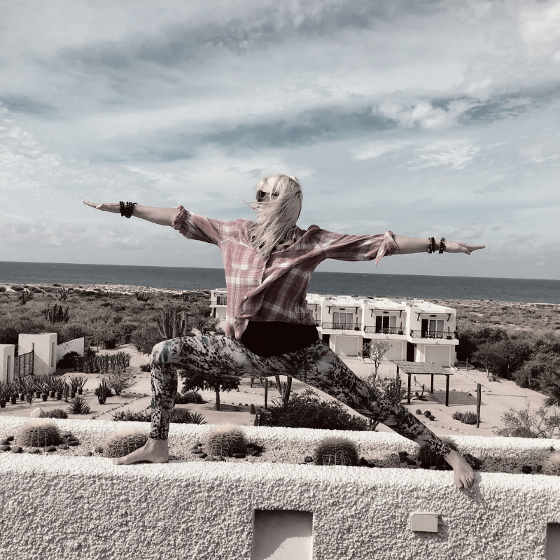 A woman with short blonde hair sitting on a beige bench, writing in a notebook with a pen, in a room with natural sunlight, a large plant, and a framed abstract art piece.