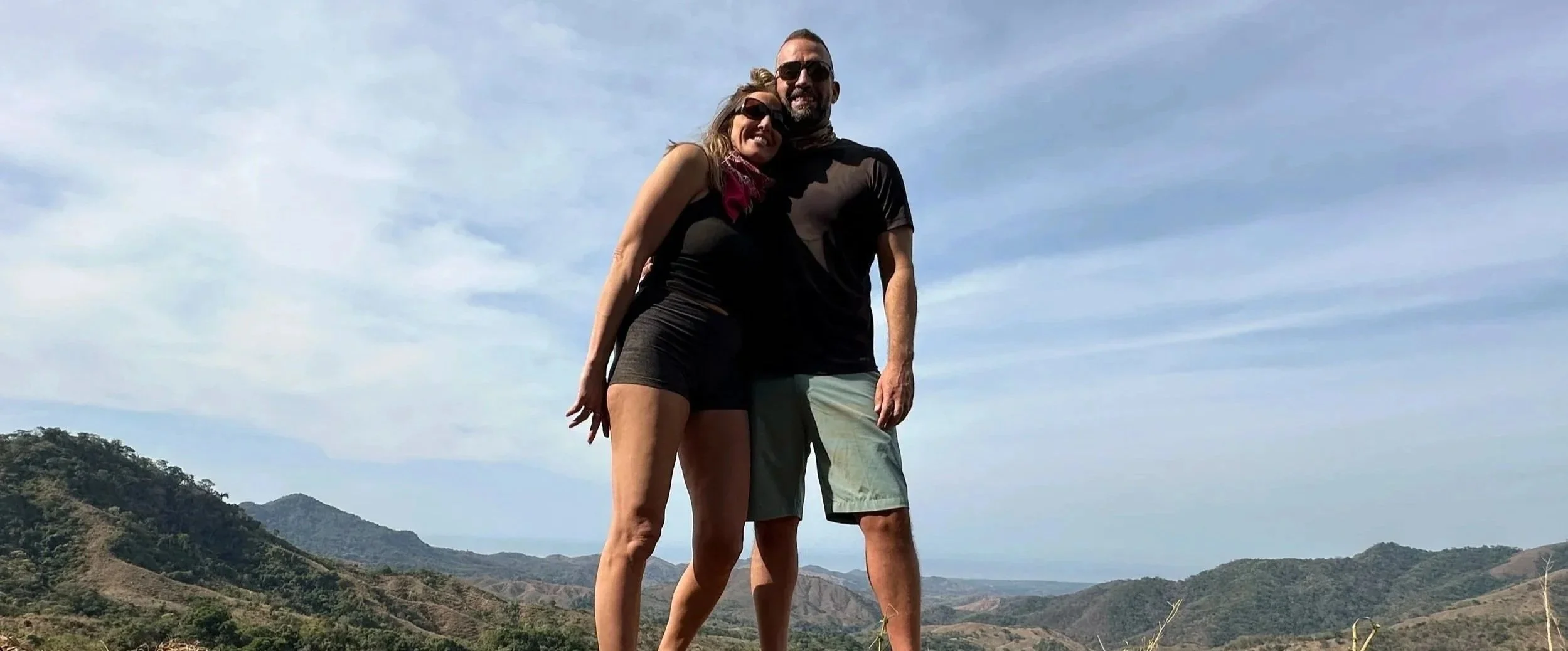A smiling couple embracing outdoors on a hill with mountains and a blue sky in the background on a Studio One Yoga Retreat