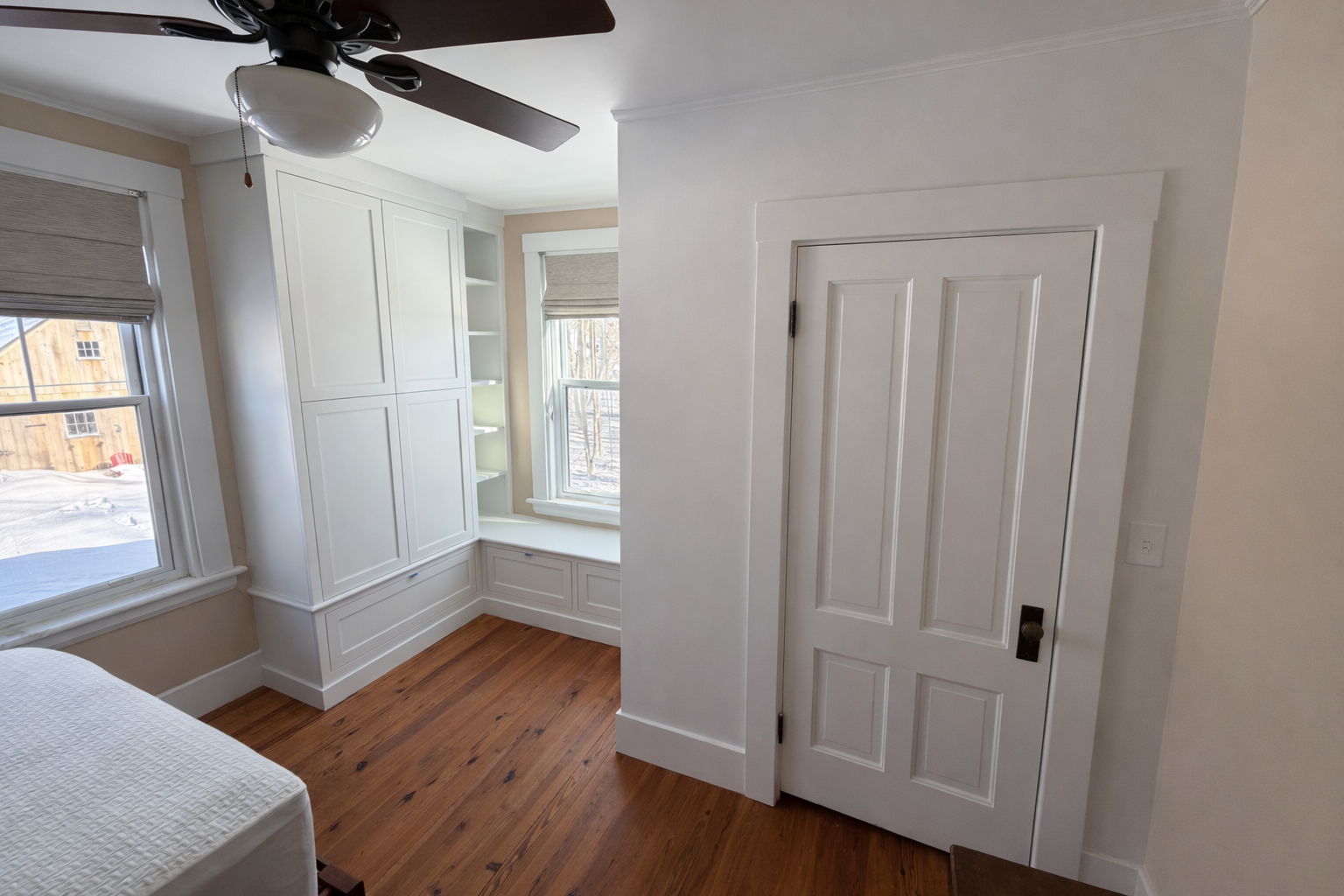 A room with white walls, hardwood flooring, two windows with blinds, a built-in white cabinet with open shelves, and a ceiling fan with lights.