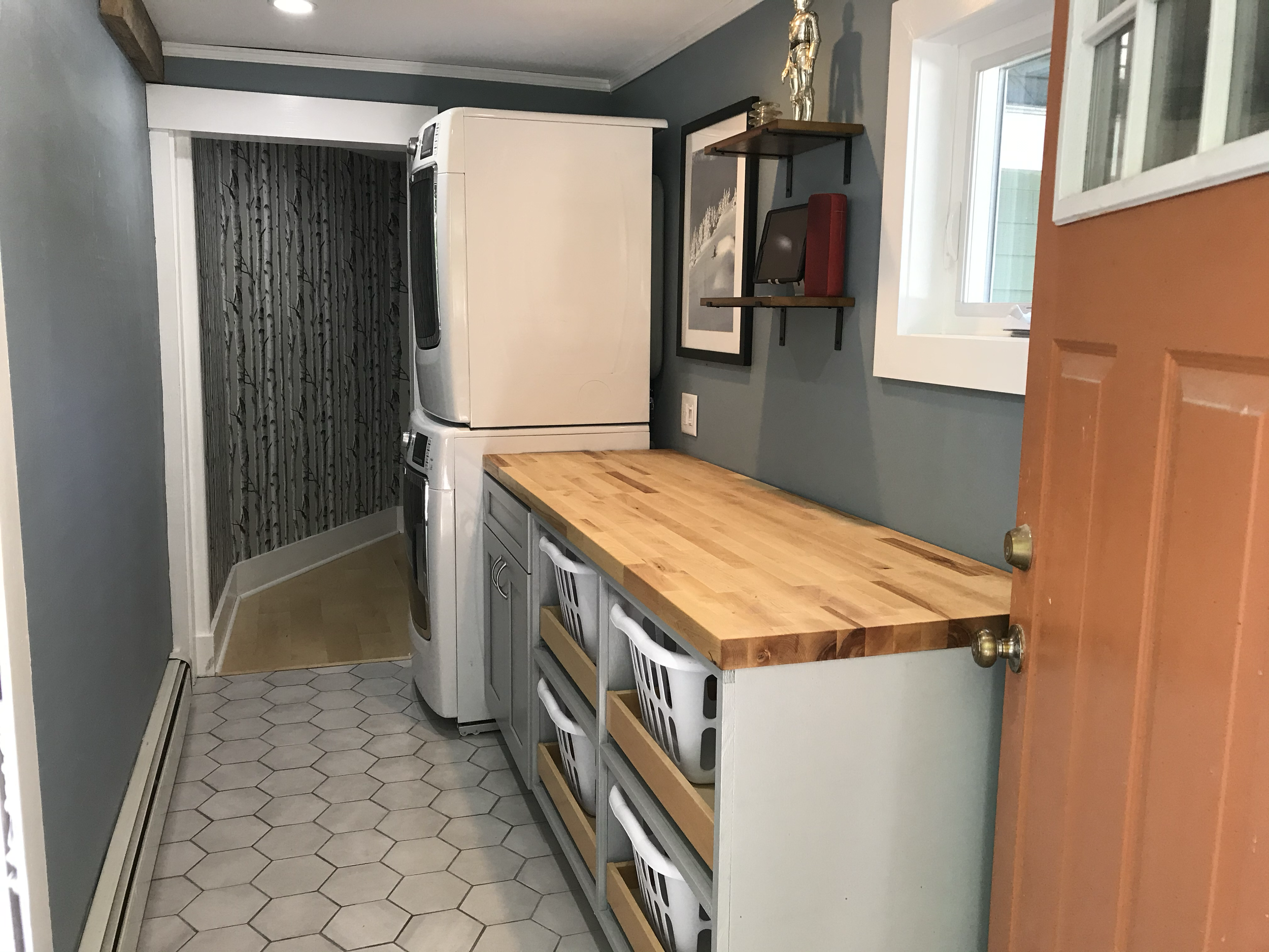 Laundry room with washing machine and dryer stacked, wooden countertop, gray cabinets, hexagon floor tiles, and wall shelves with decorative items.