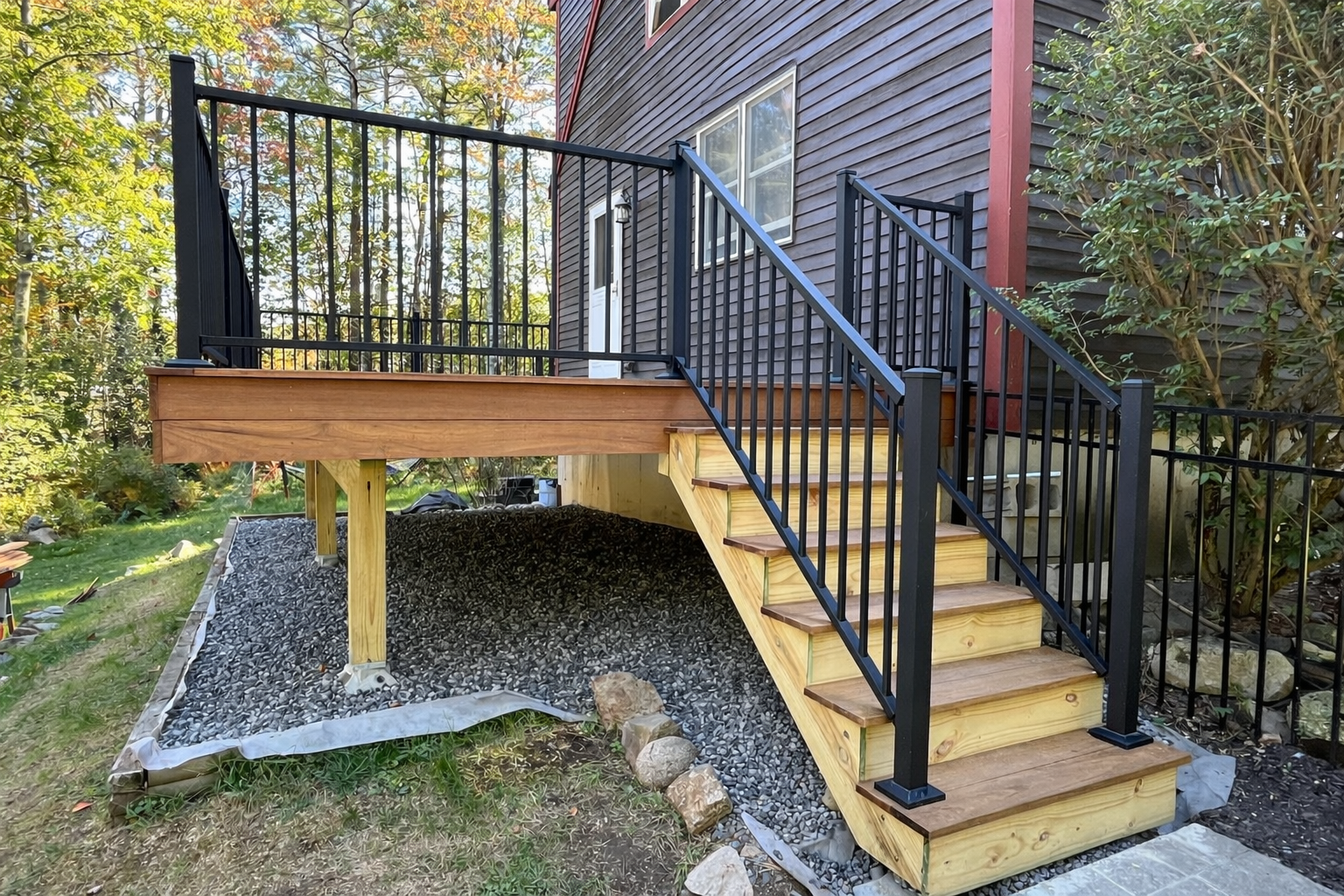 Newly built wooden deck with black metal railing and stairs attached to a house with gray siding, under construction with gravel underneath, in a backyard with trees and plants.
