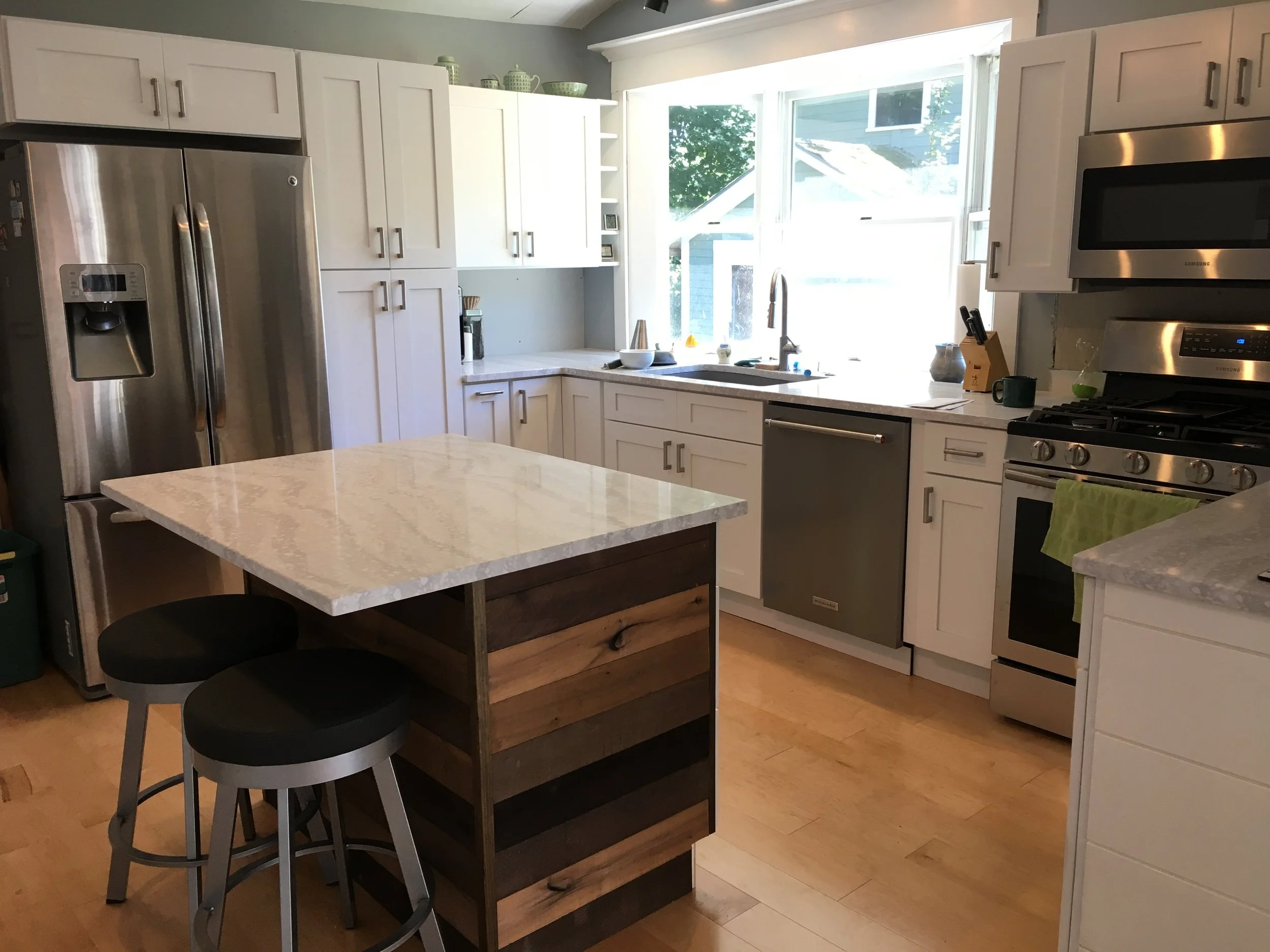 Kitchen with white cabinets, stainless steel appliances, a wooden island with a marble top, stools, and a large window.
