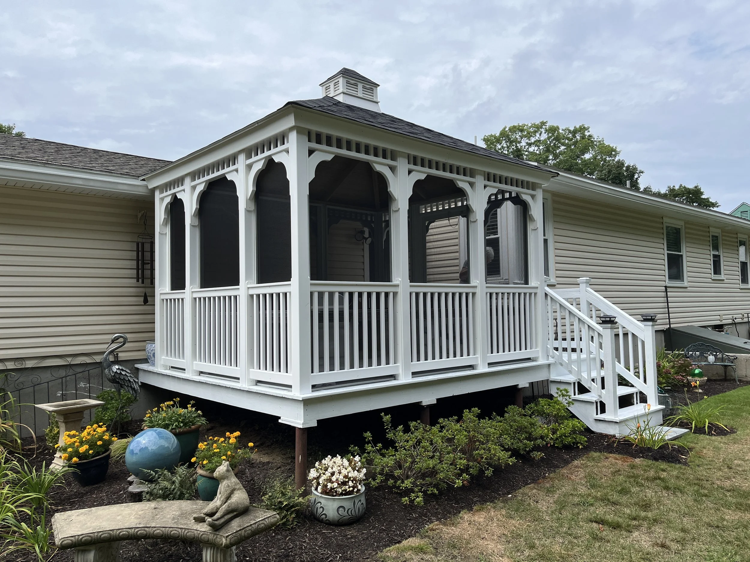 A white screened porch with stairs leading up to it, attached to a beige house with multiple windows and a sloped roof, surrounded by a garden with flowers, decorative items, and outdoor furniture.